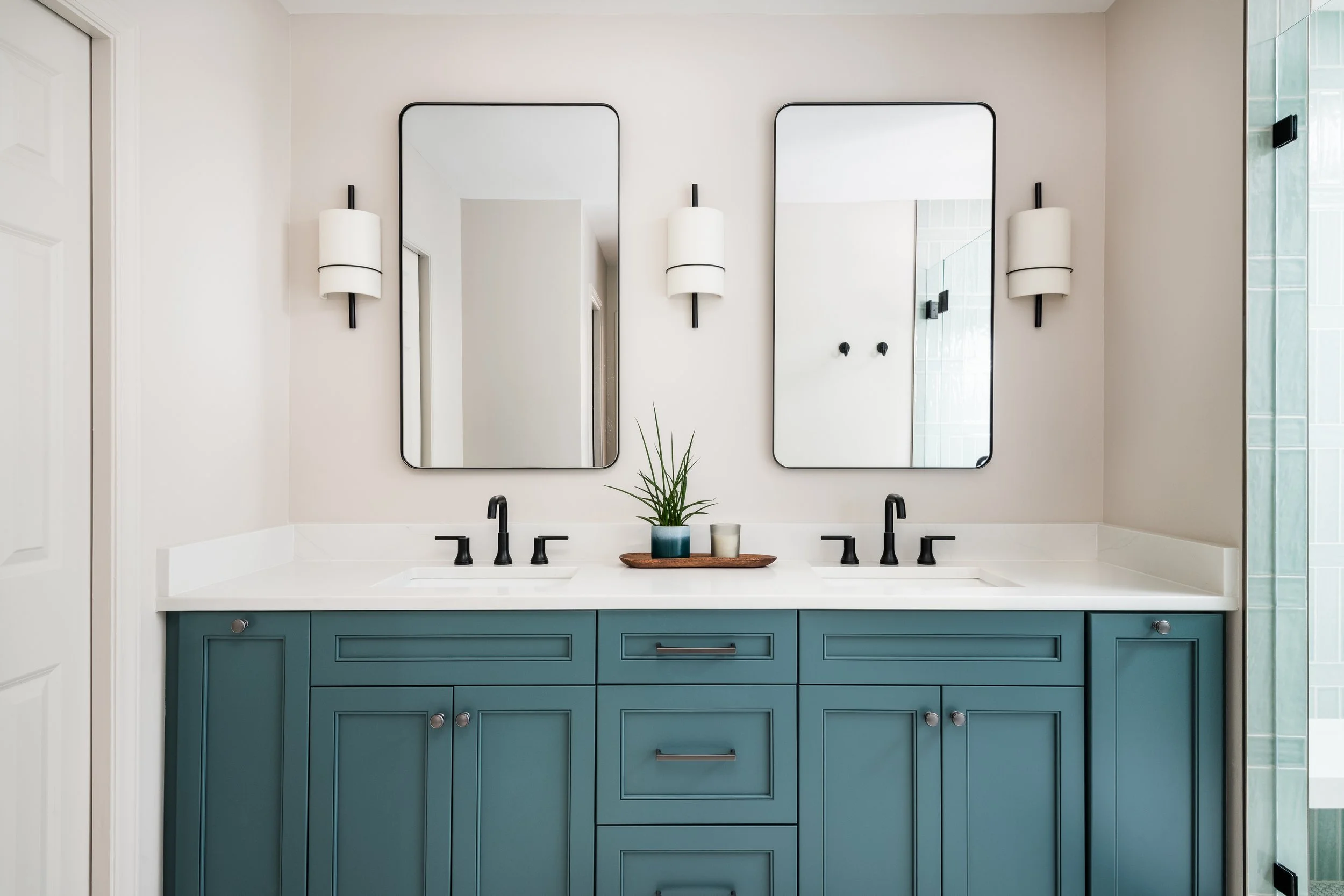 A modern bathroom vanity with a white countertop, two black faucets, and a blue cabinet with drawers. Two rectangular mirrors are mounted above, flanked by matching wall sconces. A small tray with a plant and two cups is on the counter. Part of a sho
