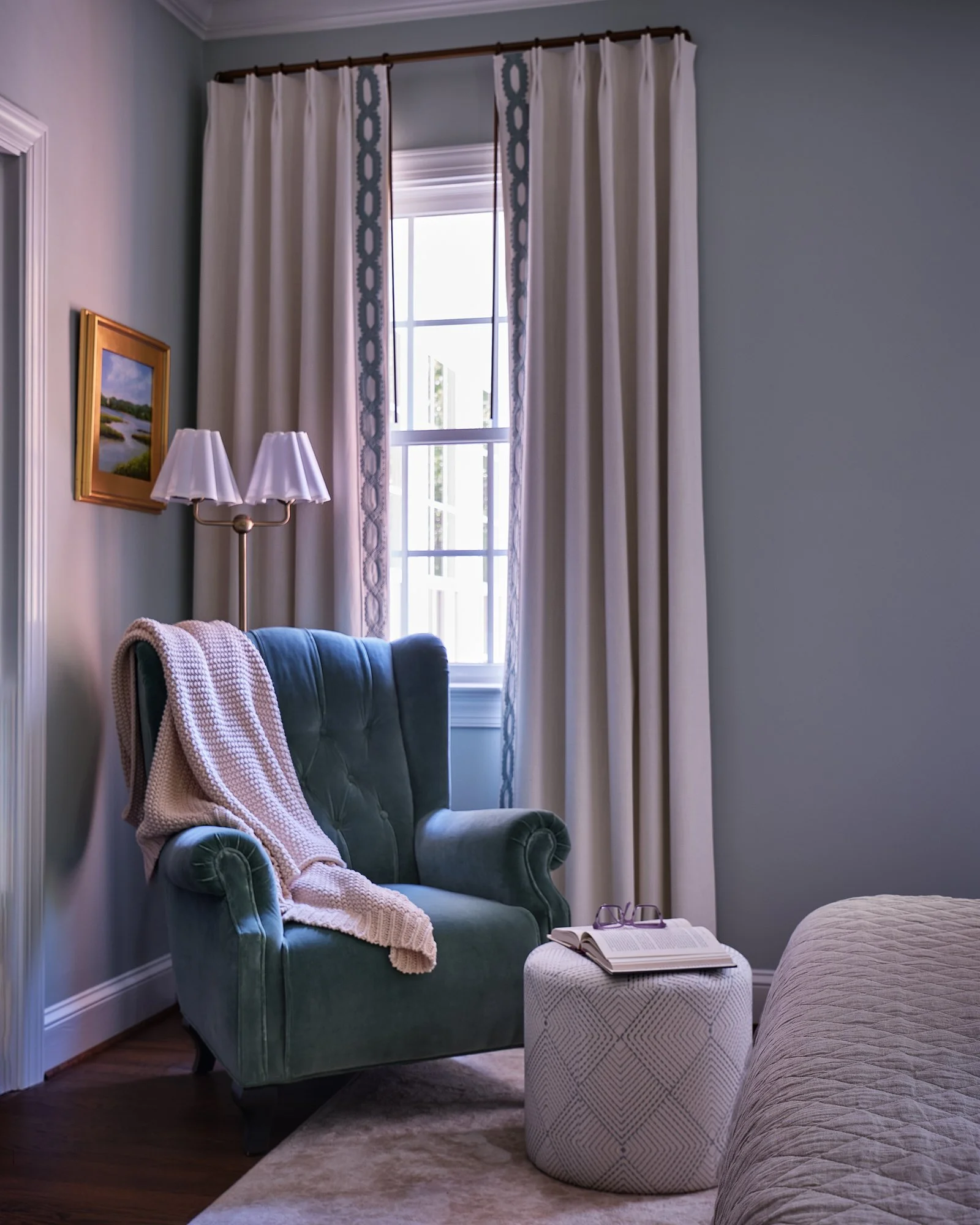 A cozy living room corner featuring a green upholstered armchair with a blanket draped over it, a small round patterned side table with an open book and reading glasses, a window with white curtains, a framed landscape painting on the wall, and a bei