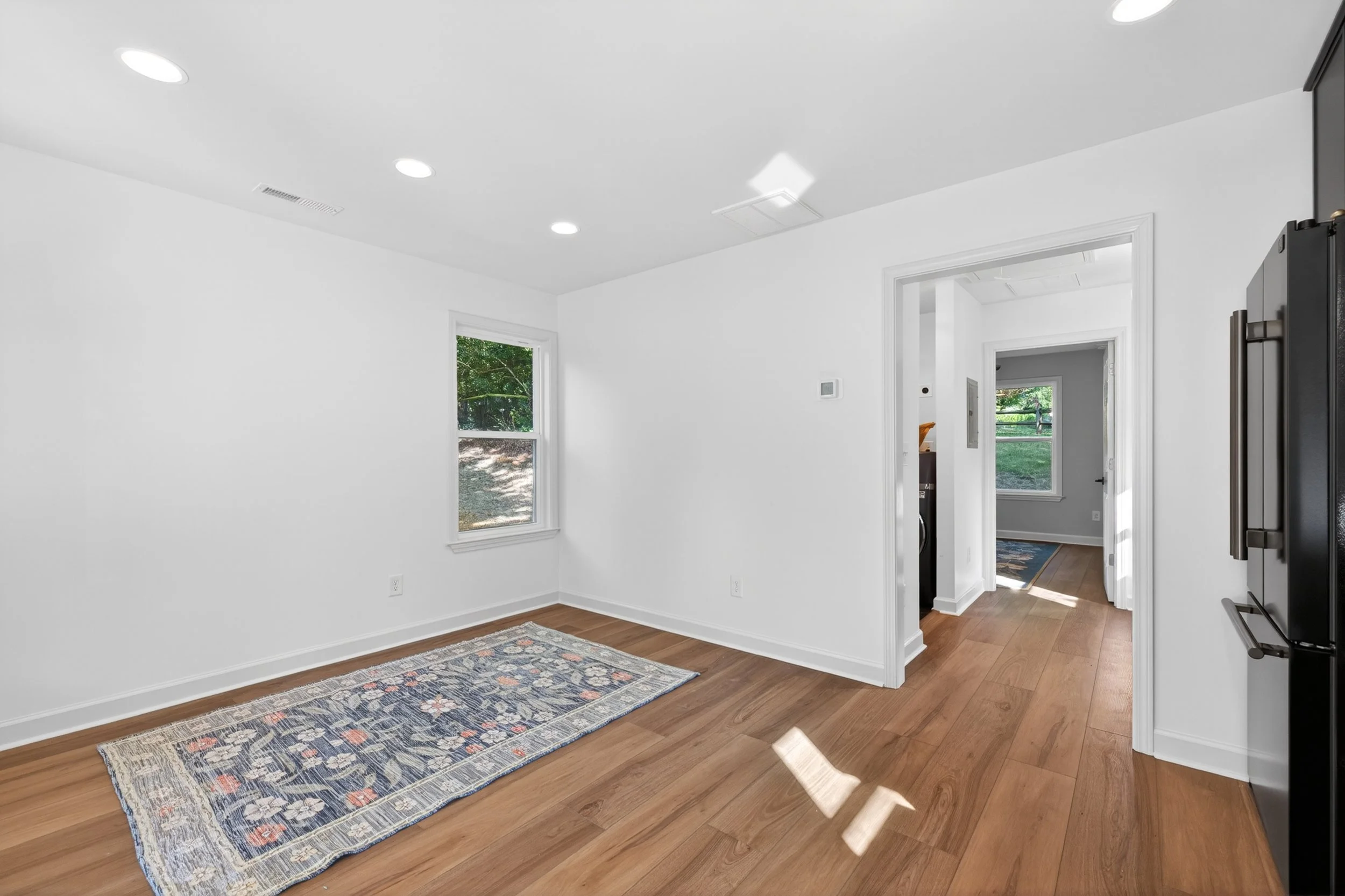 Empty white room with hardwood floors, patterned rug, two windows showing greenery outside, open doorway leading to another room, and a mounted TV on the wall.