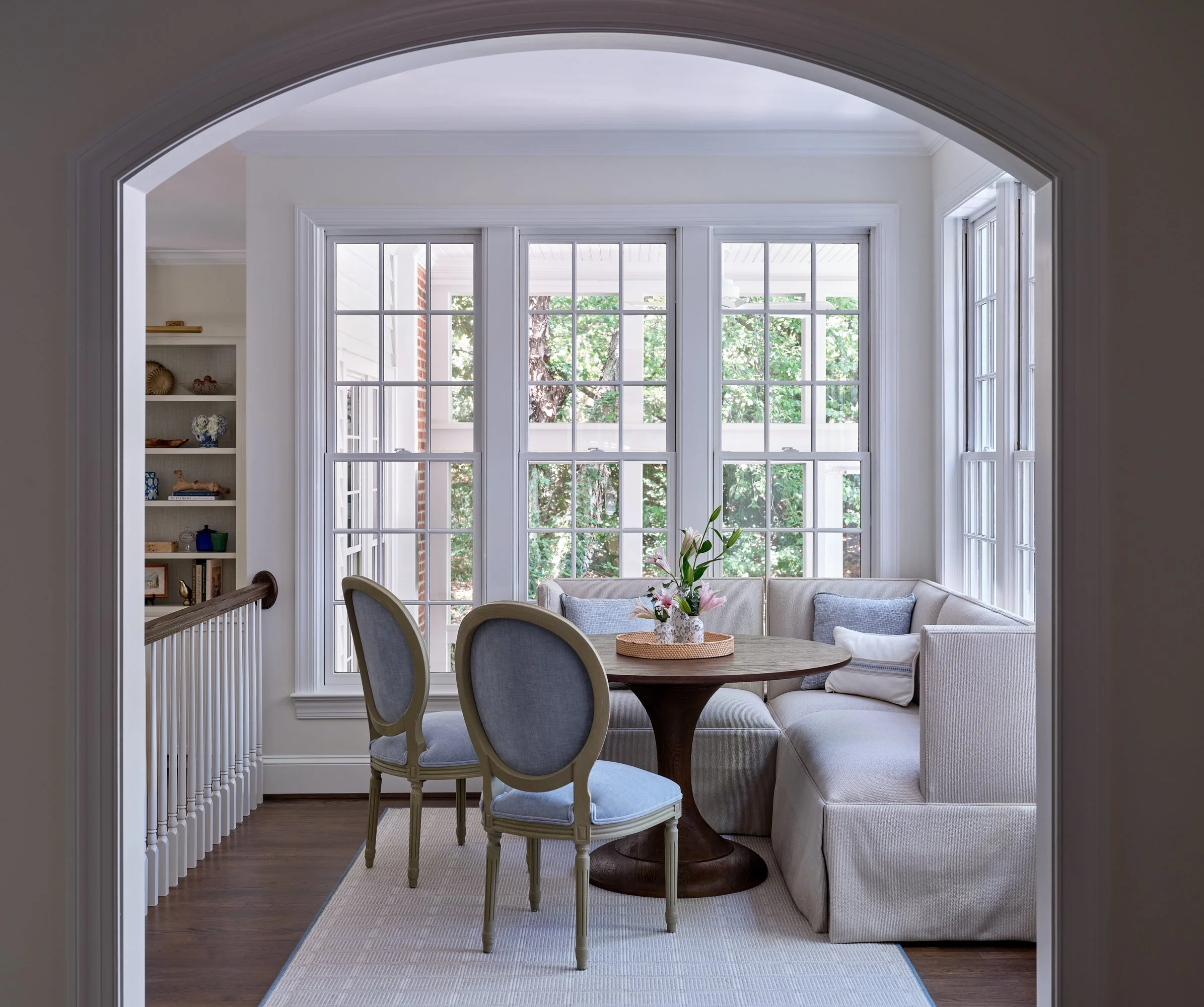 Interior of a living room with a large window showing trees outside, dining table with a flower arrangement, cream-colored chairs, a sectional sofa, and a built-in bookshelf.