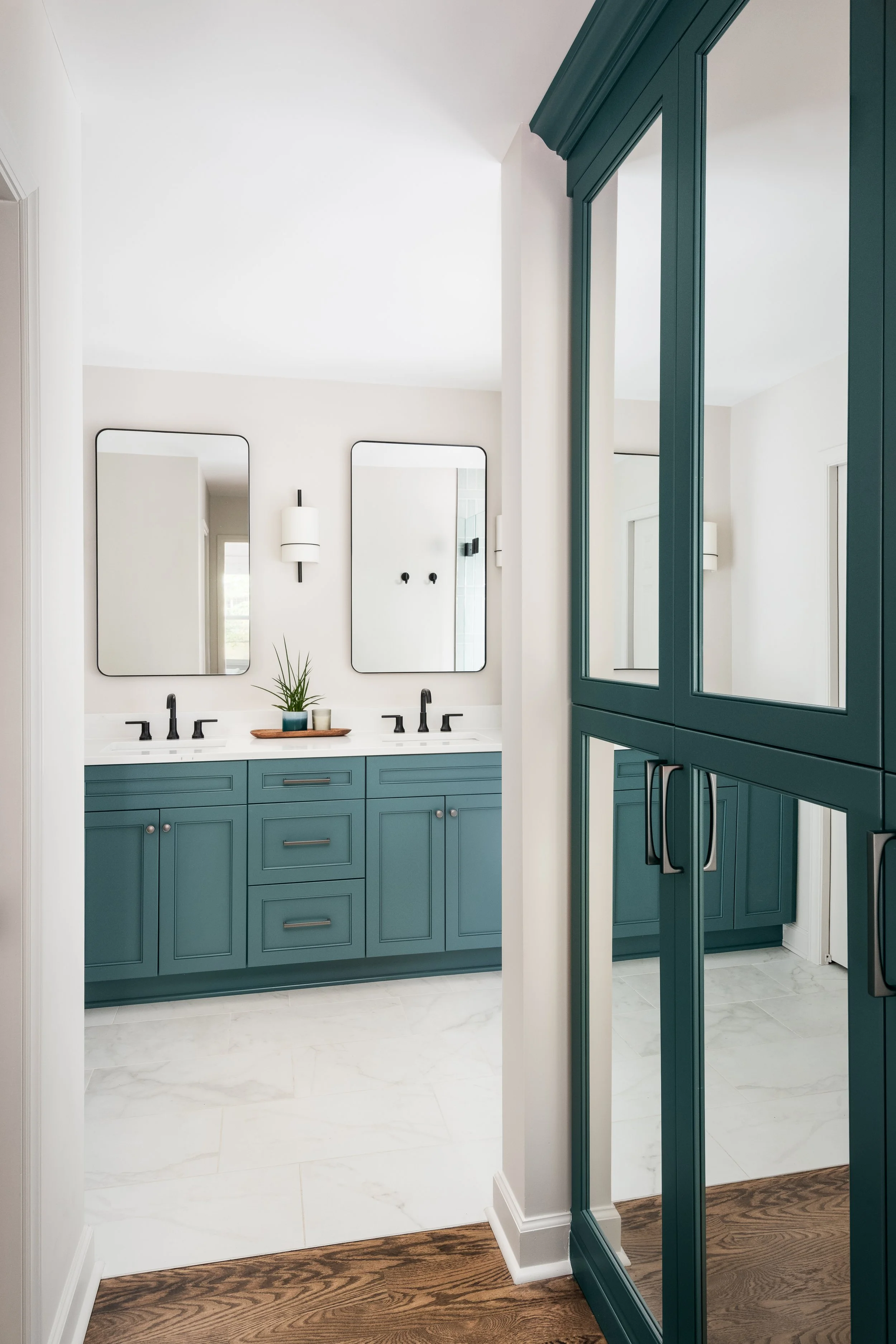 A bathroom vanity area with teal cabinetry, two mirrors, black faucets, a small plant, and a tray with cups. The room has white walls, white marble floor tiles, and additional teal cabinetry to the right.