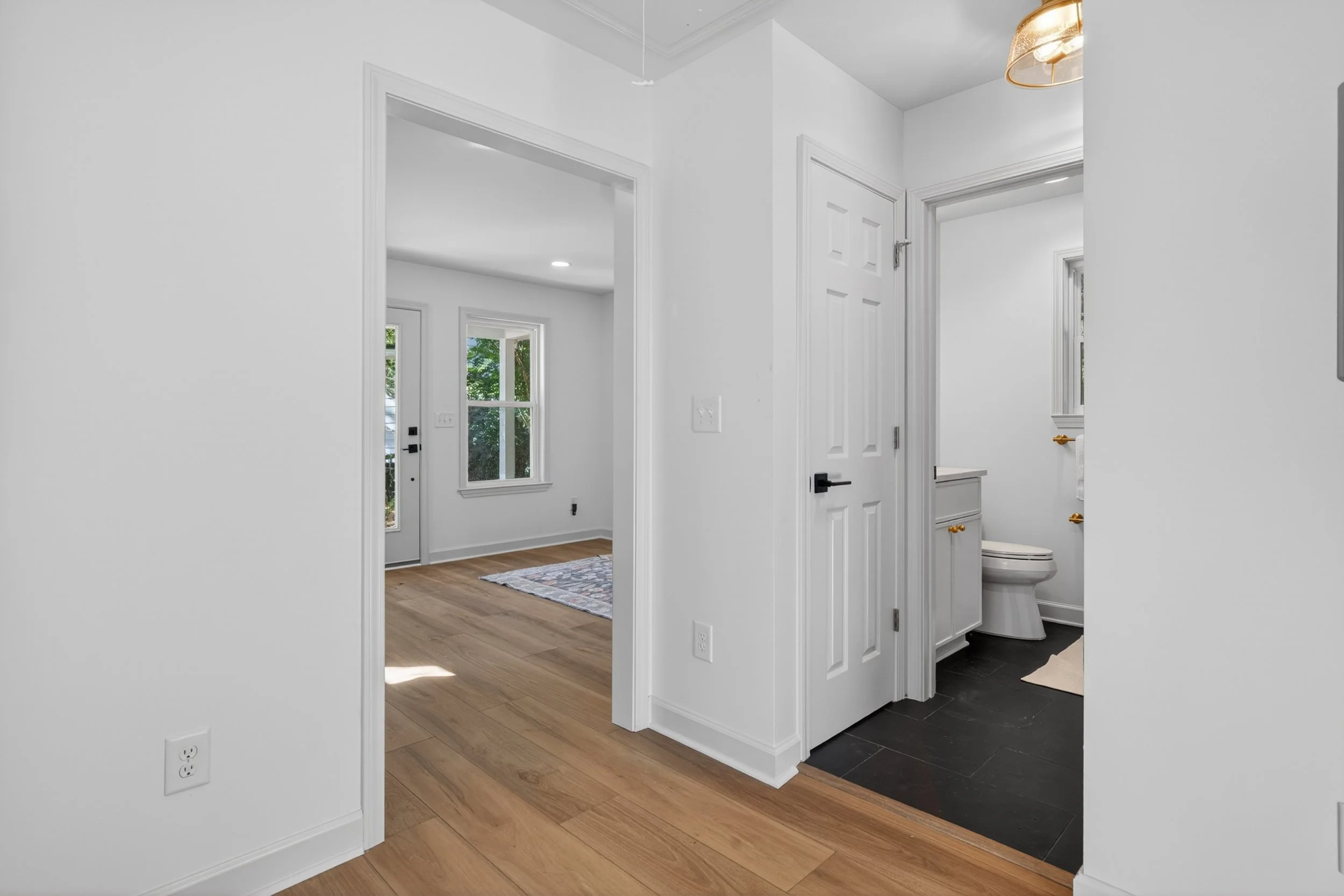 Interior view of a modern home with white walls, wood and tile flooring, showing a bathroom with a toilet and a room with a window and door.
