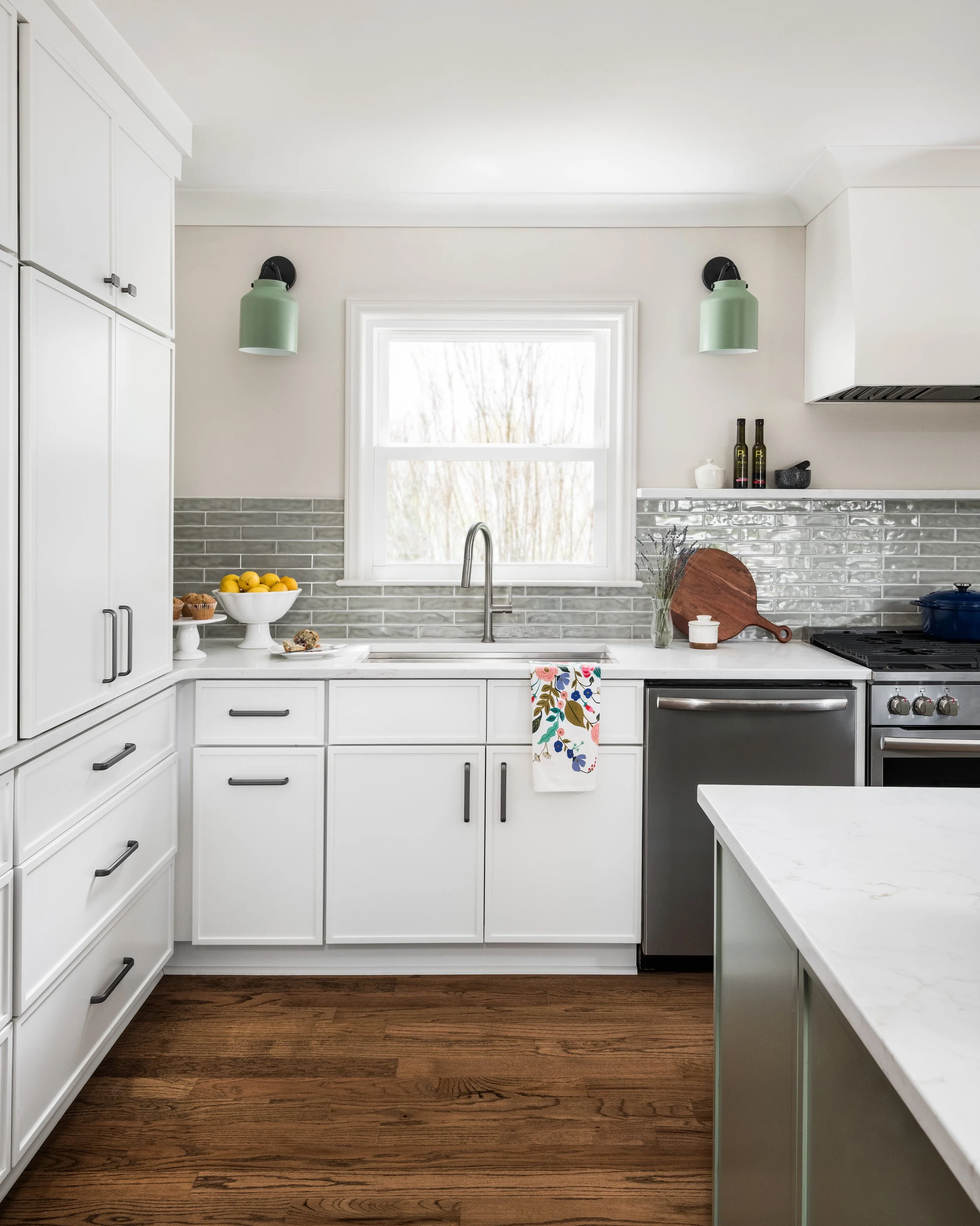 A kitchen with white cabinets, a white marble countertop, and stainless steel appliances. There are hanging green pendant lights above the sink, a bowl of lemons on the counter, and a window with a white frame and a view of trees outside.
