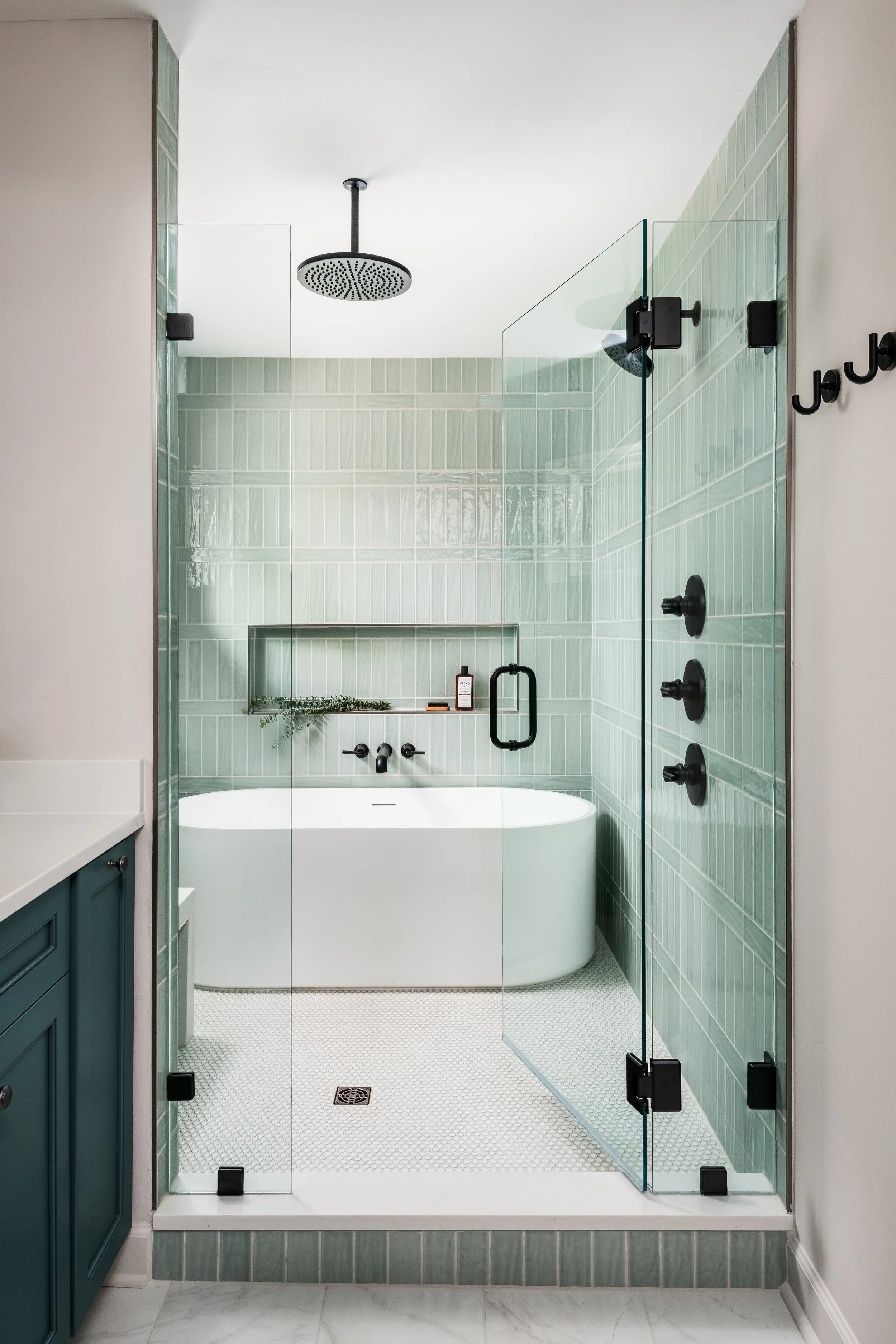 Modern bathroom with a glass-enclosed shower featuring green tiled walls and a black rainfall showerhead, a bathtub underneath, and a blue vanity on the left, all with a white tiled floor.
