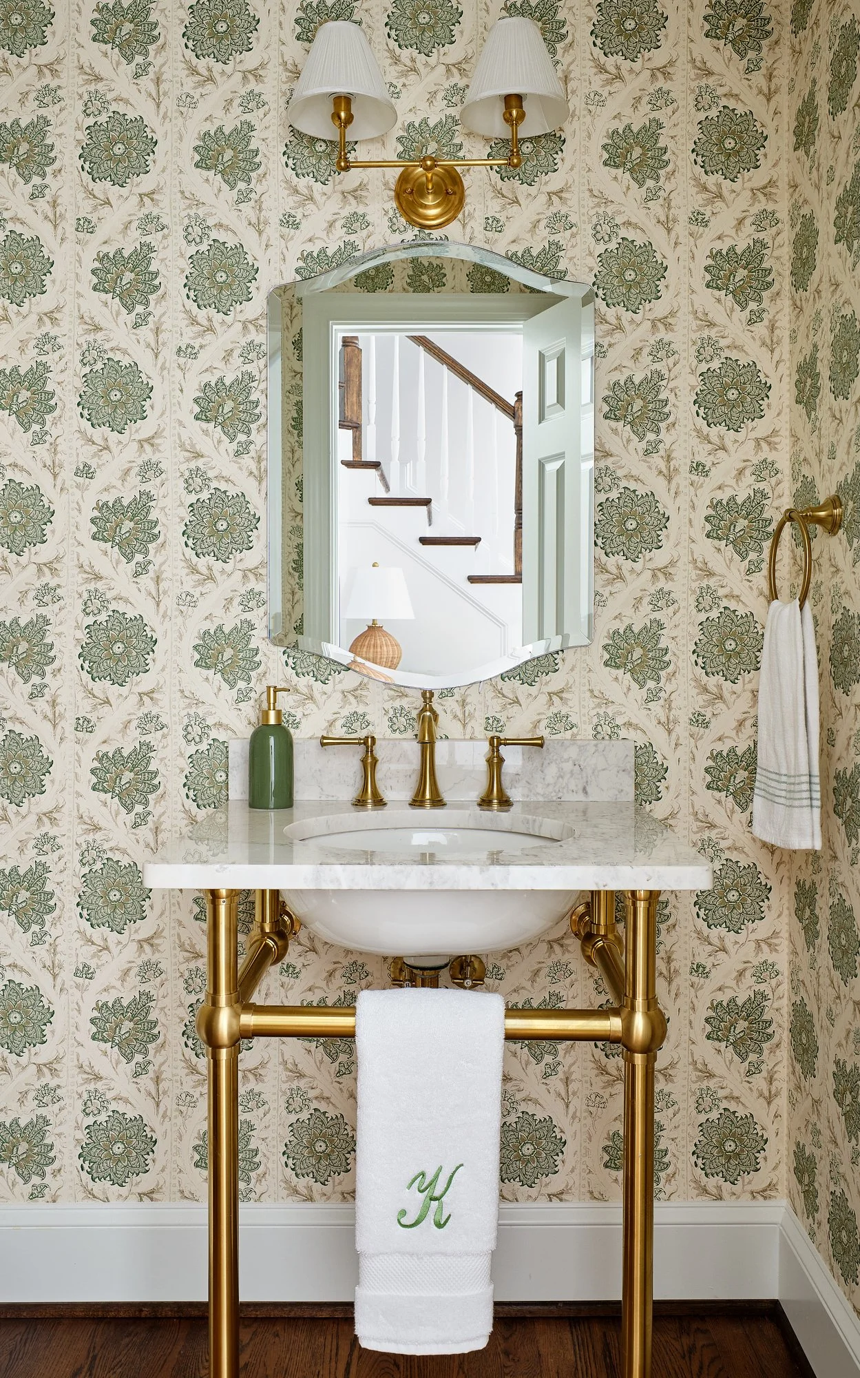 A bathroom vanity with gold plumbing, a marble countertop, a white sink, and a white towel embroidered with a green letter 'K'. A mirror is mounted above the sink, and a wall lamp with two white lampshades is above the mirror. The wall has floral pat