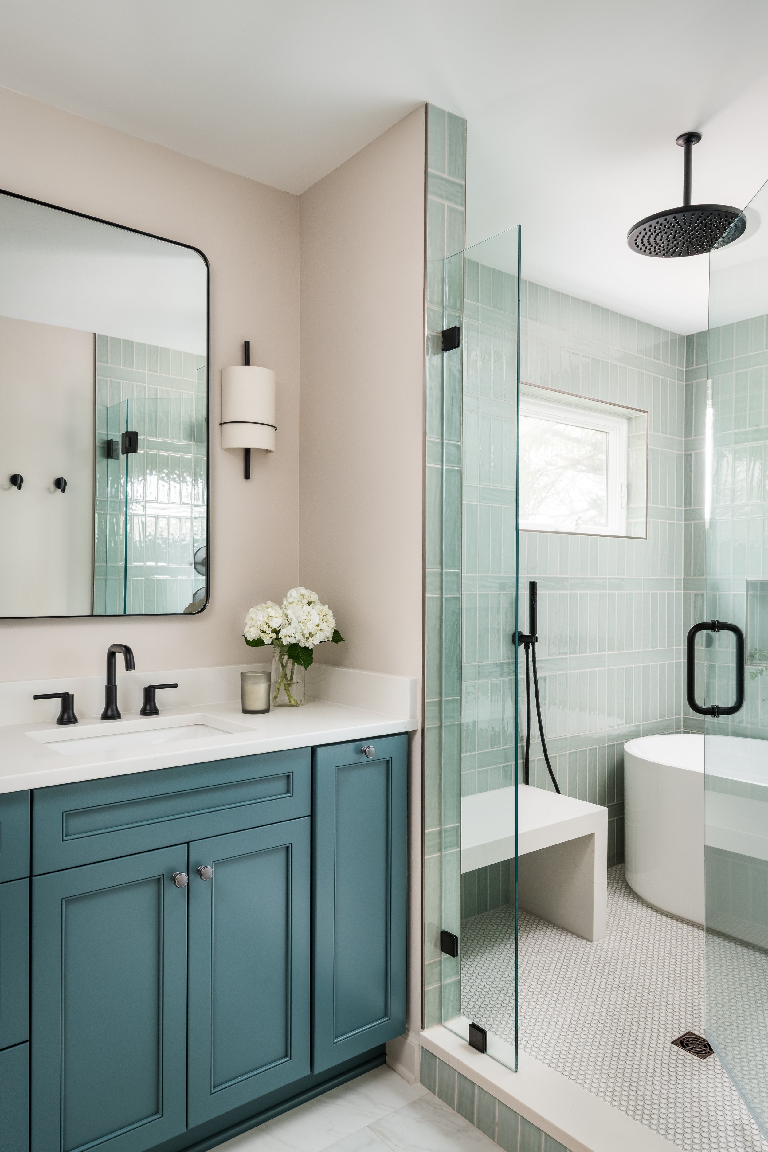 A bathroom with a vanity featuring blue cabinets, black fixtures, and a white countertop. There is a large mirror and a wall-mounted light fixture. Next to the vanity is an open shower area with green tile walls, a large black showerhead, a glass doo
