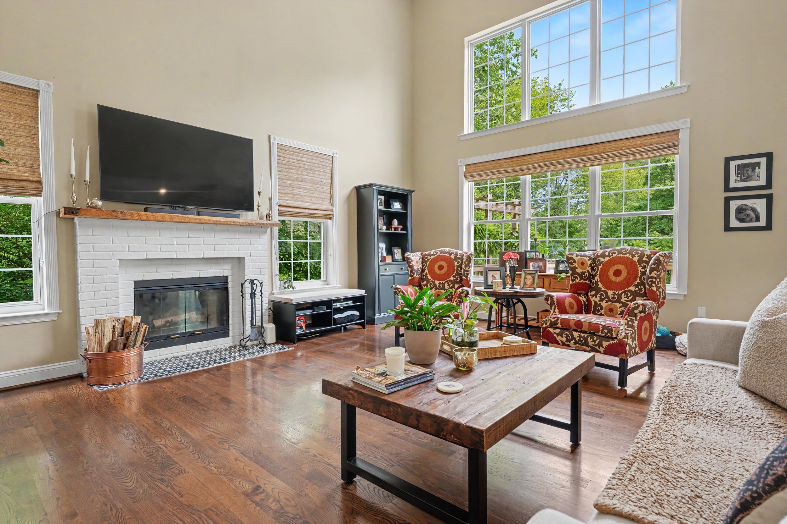 A living room with large windows, a white brick fireplace, a flat-screen TV mounted above the fireplace, a black bookshelf, a wooden coffee table with decorations, and patterned armchairs.