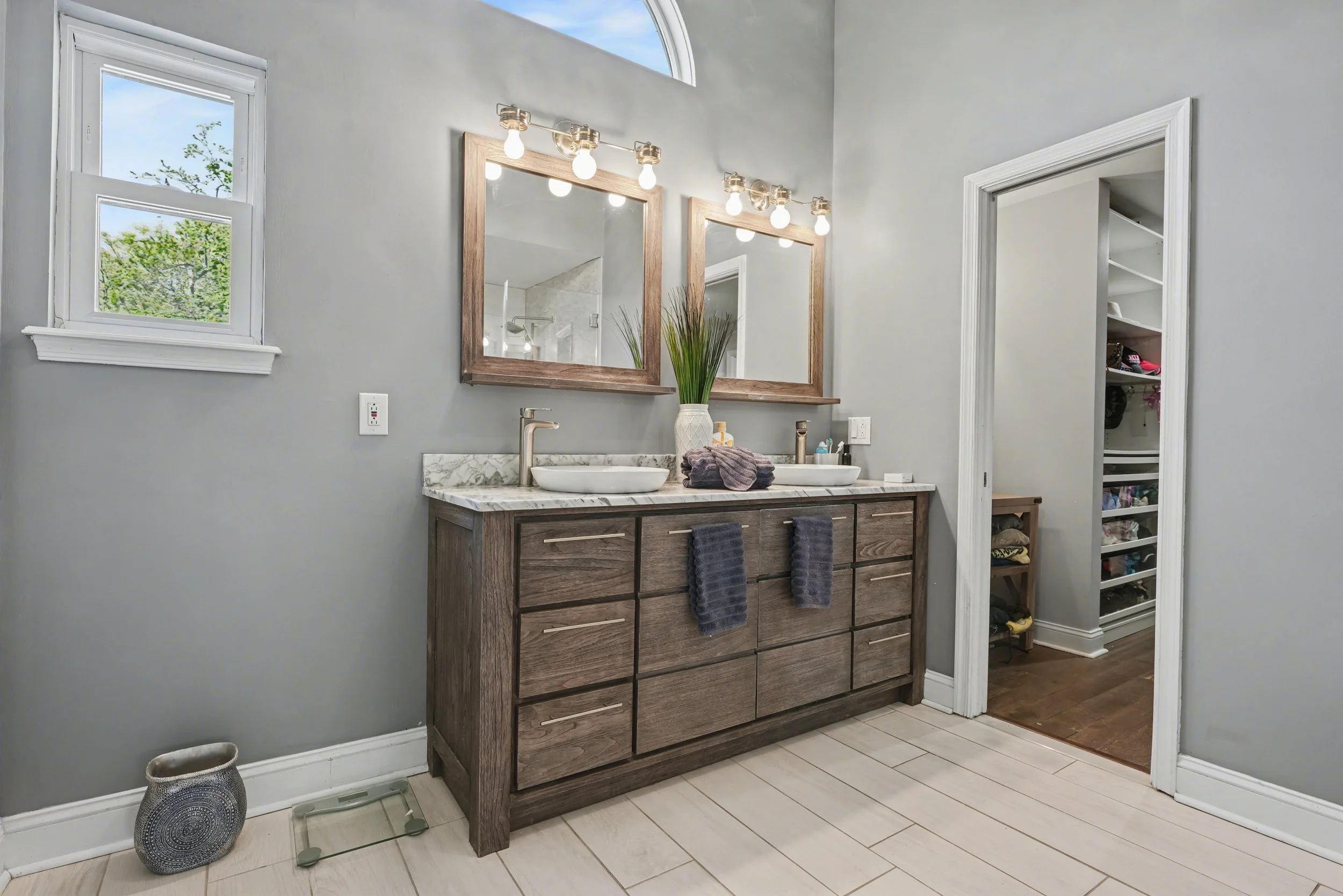 A bathroom featuring a wood vanity with double sinks, two mirrors with wooden frames, and a vase with tall grass on the countertop. There are three light bulbs above each mirror. A window and an arched window are visible on the wall, showing greenery