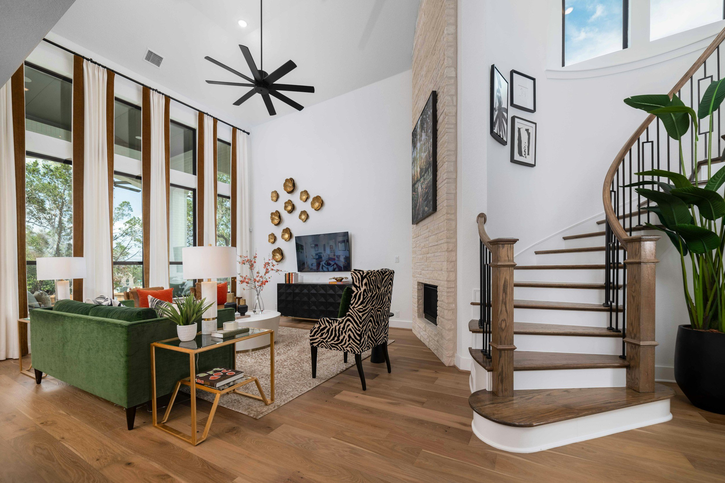 Open living room with large windows, beige curtains, wood flooring, a black ceiling fan, a white brick fireplace, various wall art, a green sofa, zebra-print chair, and a staircase with wooden handrail and black metal balusters.