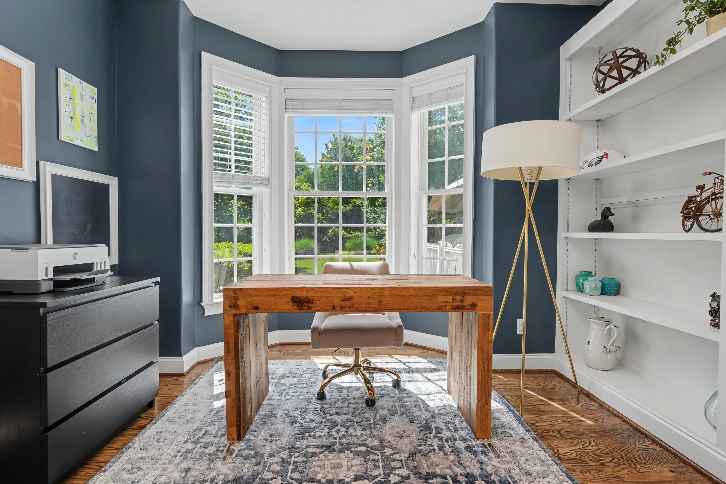 A cozy home office space with large bay window showcasing greenery outside. The room has dark blue walls, white trim, and a wooden desk with an upholstered chair. There is a black dresser with a printer, a white bookshelf with decorative items, and a