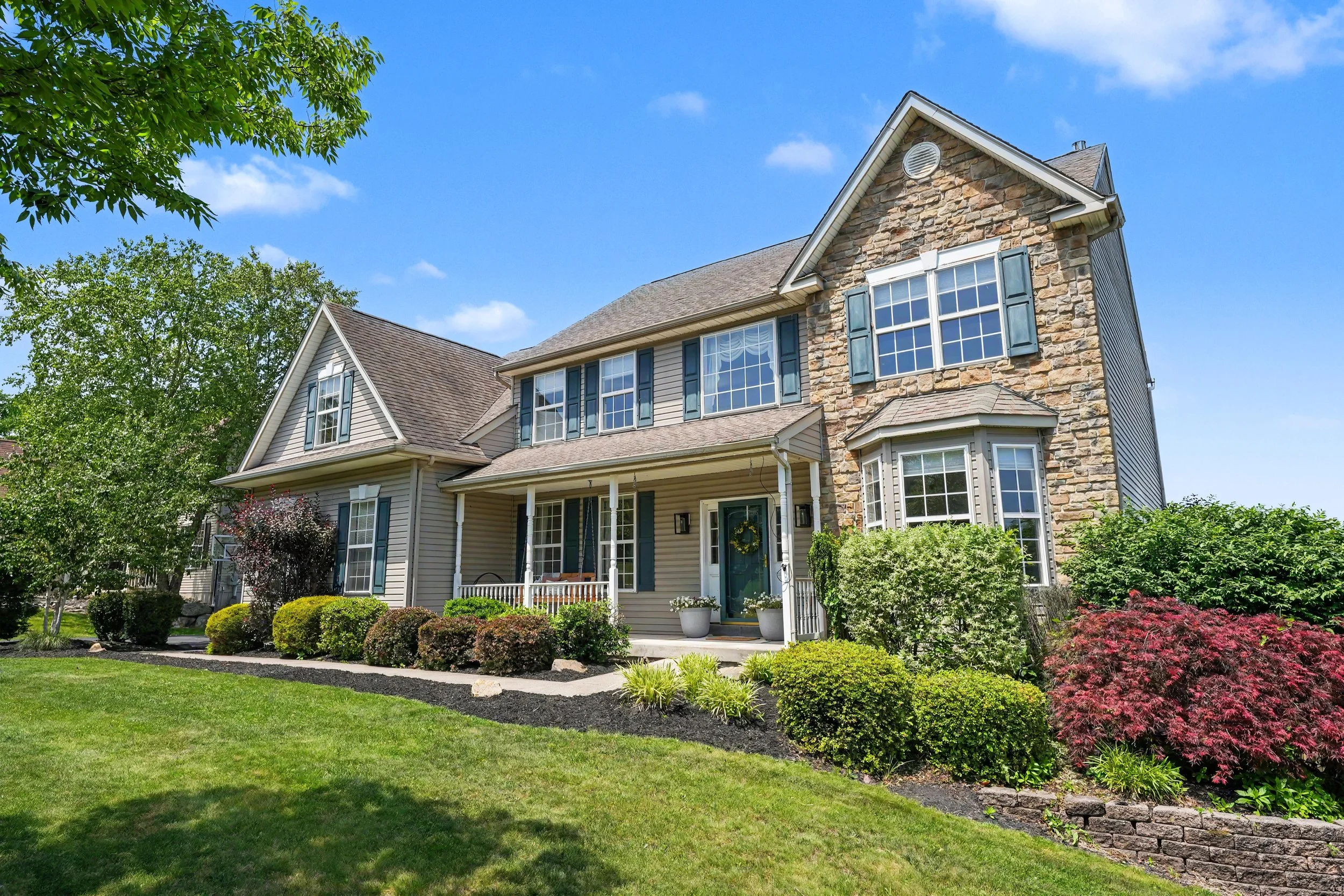 A two-story house with a stone and siding exterior, multiple large windows with shutters, a front porch with potted plants, and a well-maintained front yard with lush green grass and colorful shrubs under a bright blue sky.