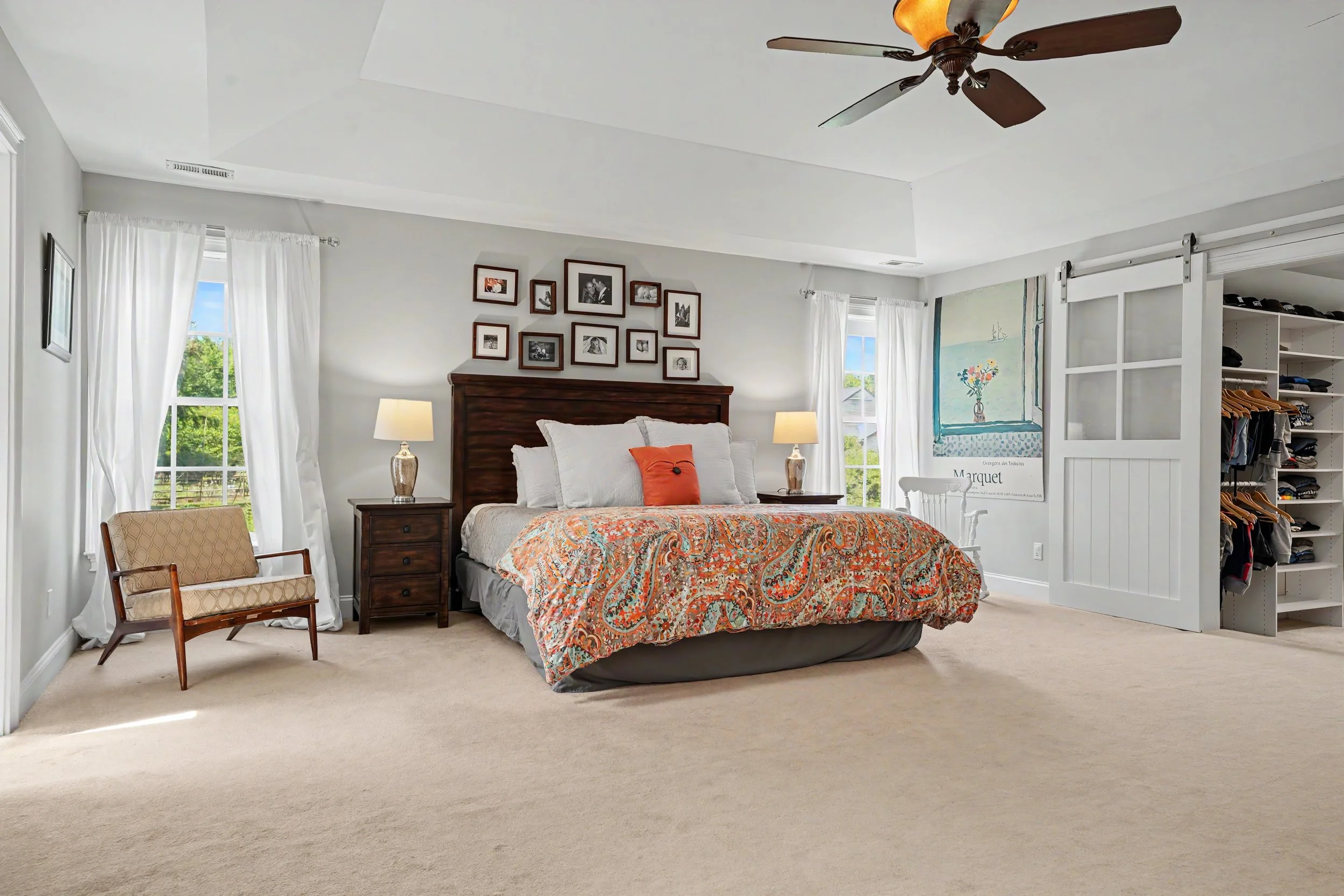 A bedroom with a large bed covered in a patterned orange and cream quilt, two nightstands with lamps, a picture gallery above the bed, a wooden armchair, and a white sliding barn door closet revealing shelves with clothes. The room has windows with w