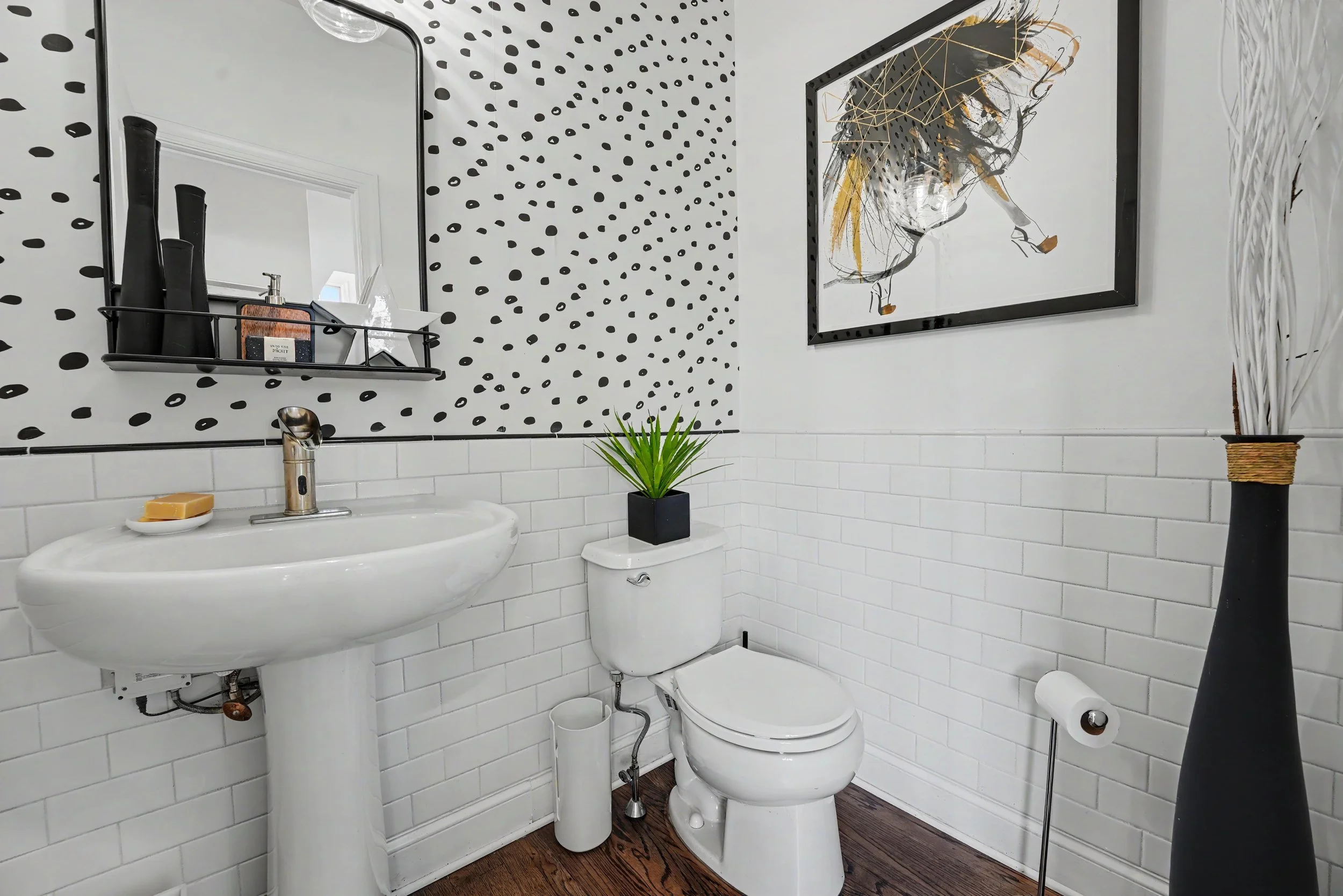 A small bathroom with white subway tile on the walls, black accents, a framed abstract art piece, a potted plant, a soap dish, and toiletries on a black wall shelf above the sink.