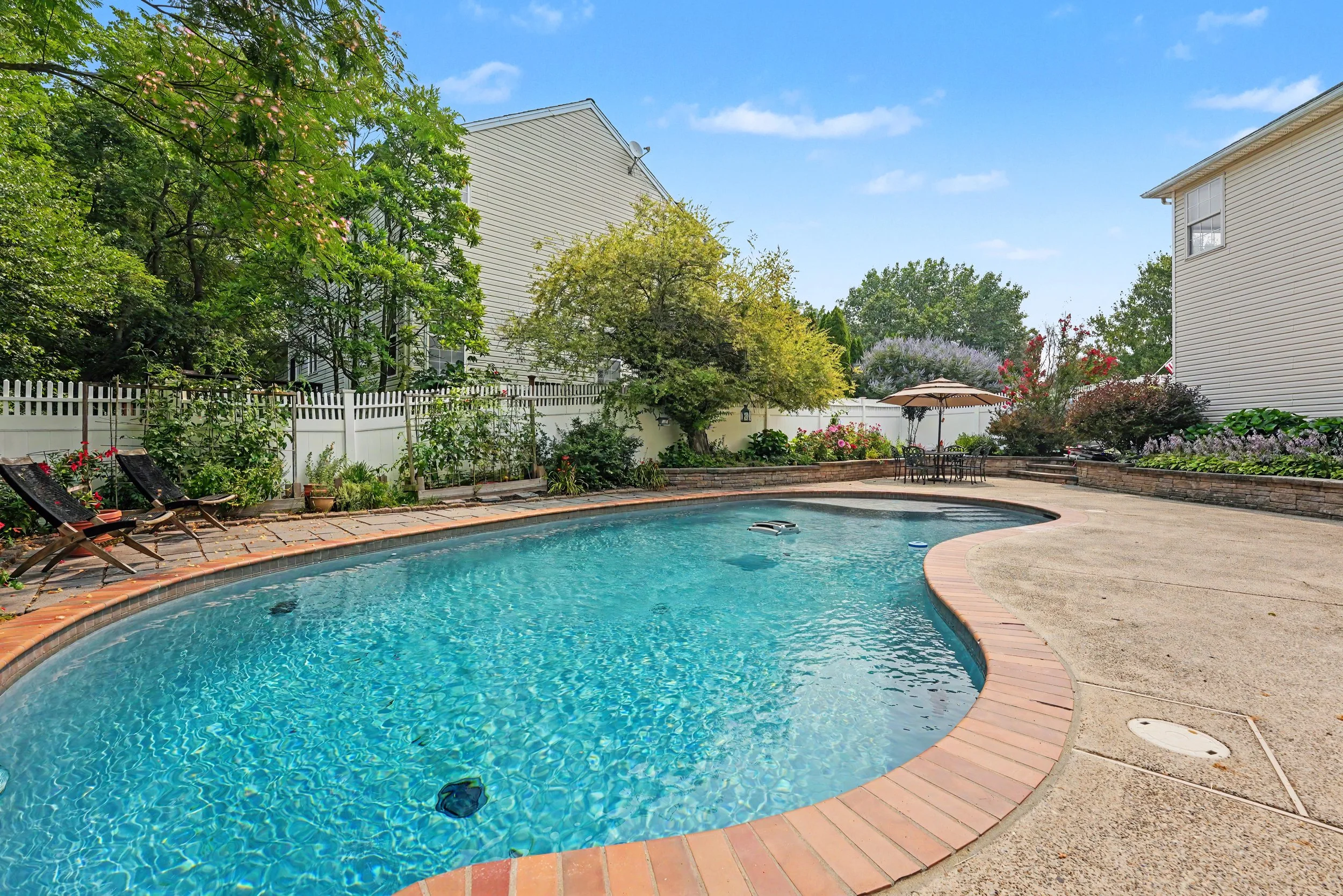 A backyard with a swimming pool surrounded by a paved deck, green trees, bushes, and flowers, with white fence, patio furniture, and a two-story house with beige siding.