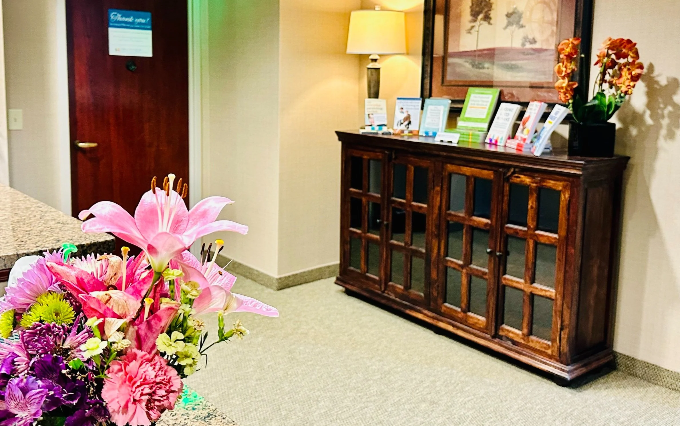 A hotel or office reception area with a bouquet of pink lilies and other flowers on a counter in the foreground, a wooden cabinet with informational brochures and a potted orchid on top, a framed picture on the wall, and a lamp providing warm lighting.