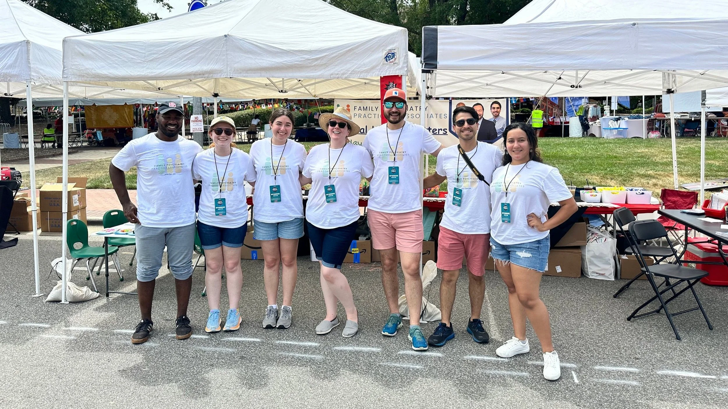 A group of seven people standing in front of a booth at an outdoor event, wearing matching white T-shirts with a logo, smiling, with tents and tables behind them.