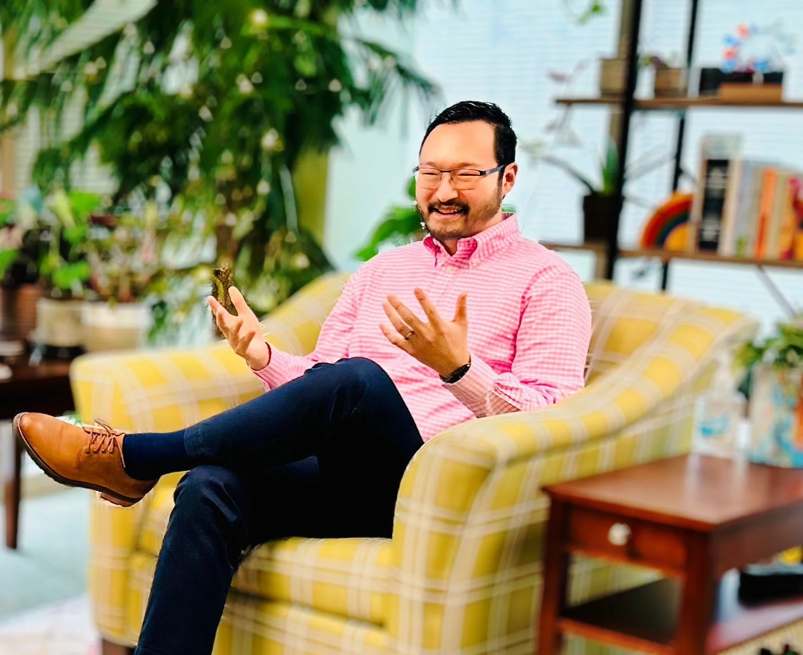 A man with glasses and a beard, wearing a pink checkered shirt, sitting on a yellow plaid sofa with crossed legs, smiling and gesturing with his hands in a cozy room filled with plants and books.