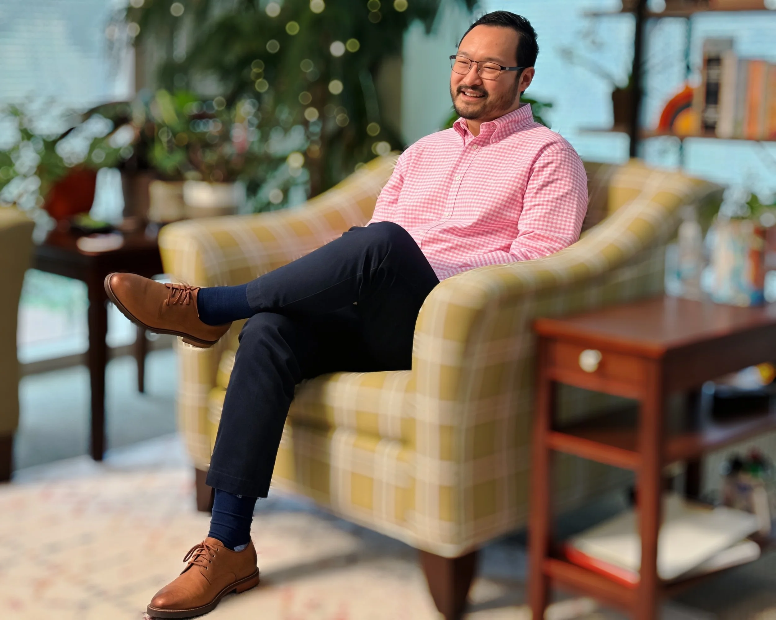 A man with glasses and a beard, wearing a pink checkered shirt, sitting on a yellow plaid sofa with crossed legs, smiling and gesturing with his hands in a cozy room filled with plants and books.