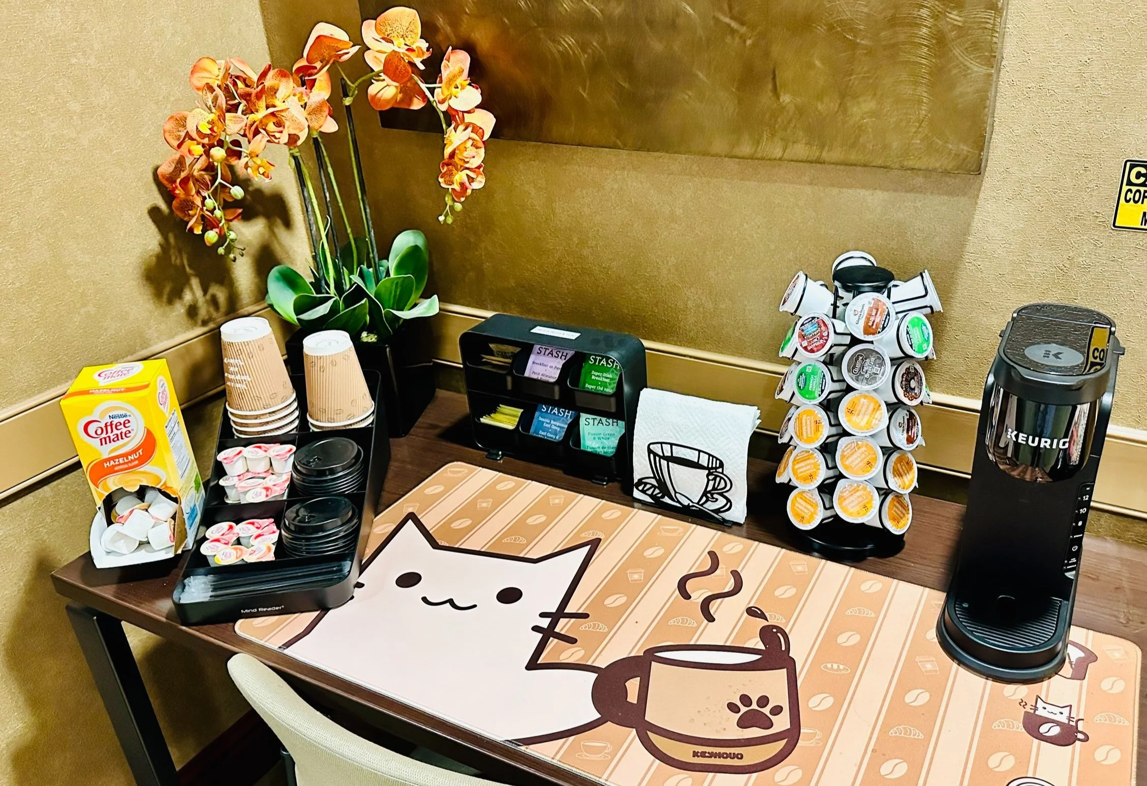 Coffee station with a cat-themed tablecloth, a potted plant with orange orchids, coffee pods, cups, napkins, a Keurig machine, and various coffee-related containers.