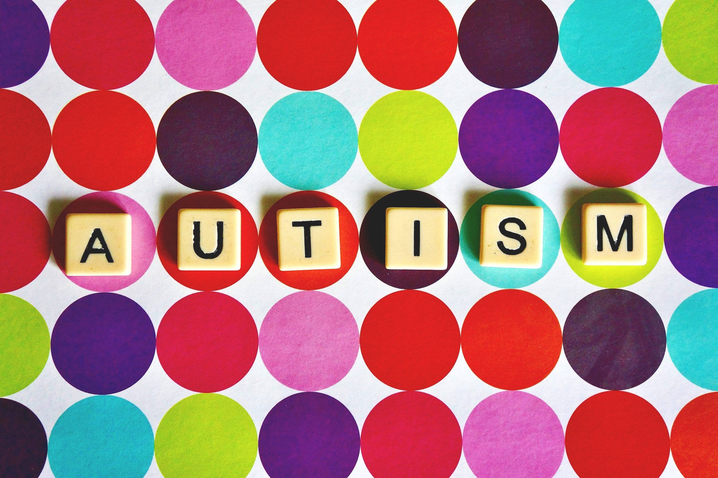 Colorful background with red, purple, blue, green, and pink dots, with cream-colored Scrabble tiles spelling out "AUTISM" in the center.