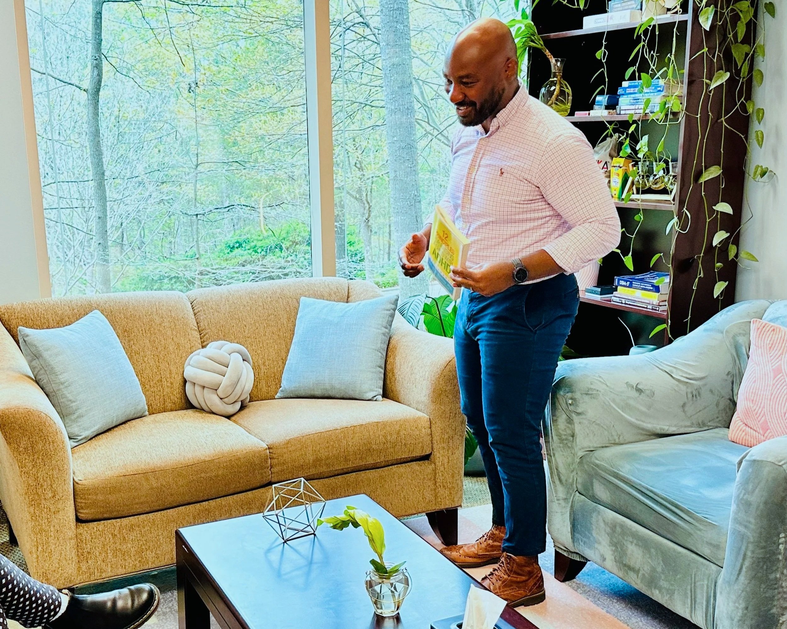 A man standing in a brightly lit living room, holding a book, with a beige sofa, a light blue velvet sofa, a coffee table with a small plant, and a bookshelf in the background. Large window with trees outside.