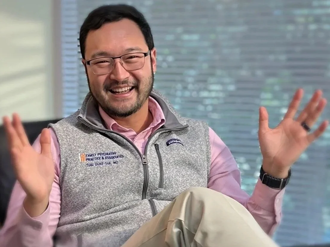 A man with glasses and a beard, wearing a pink checkered shirt, sitting on a yellow plaid sofa with crossed legs, smiling and gesturing with his hands in a cozy room filled with plants and books.