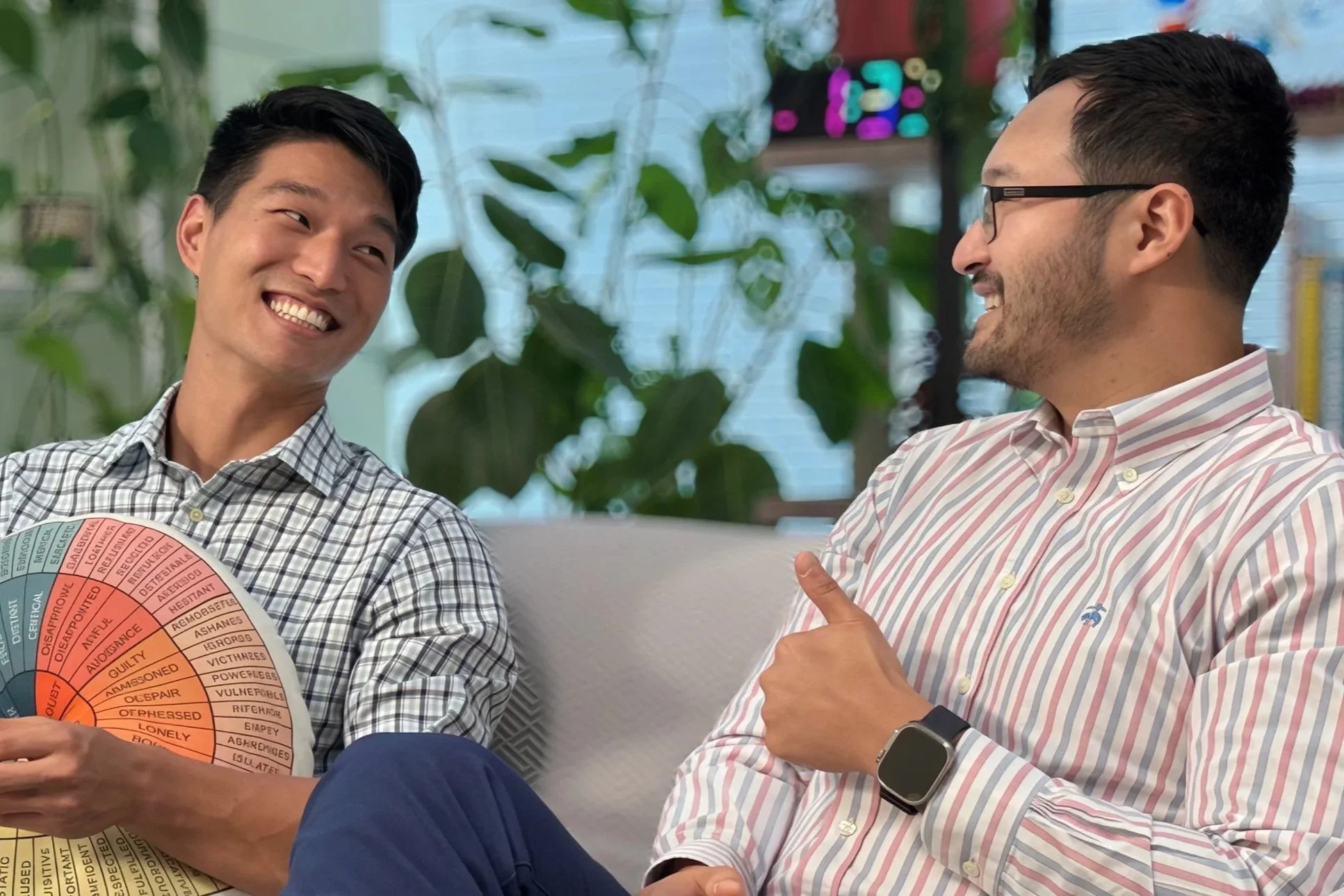 A smiling man with a bald head and beard, wearing a pink checkered shirt and dark blue jeans, sitting on a chair in a bright room, engaged in conversation.