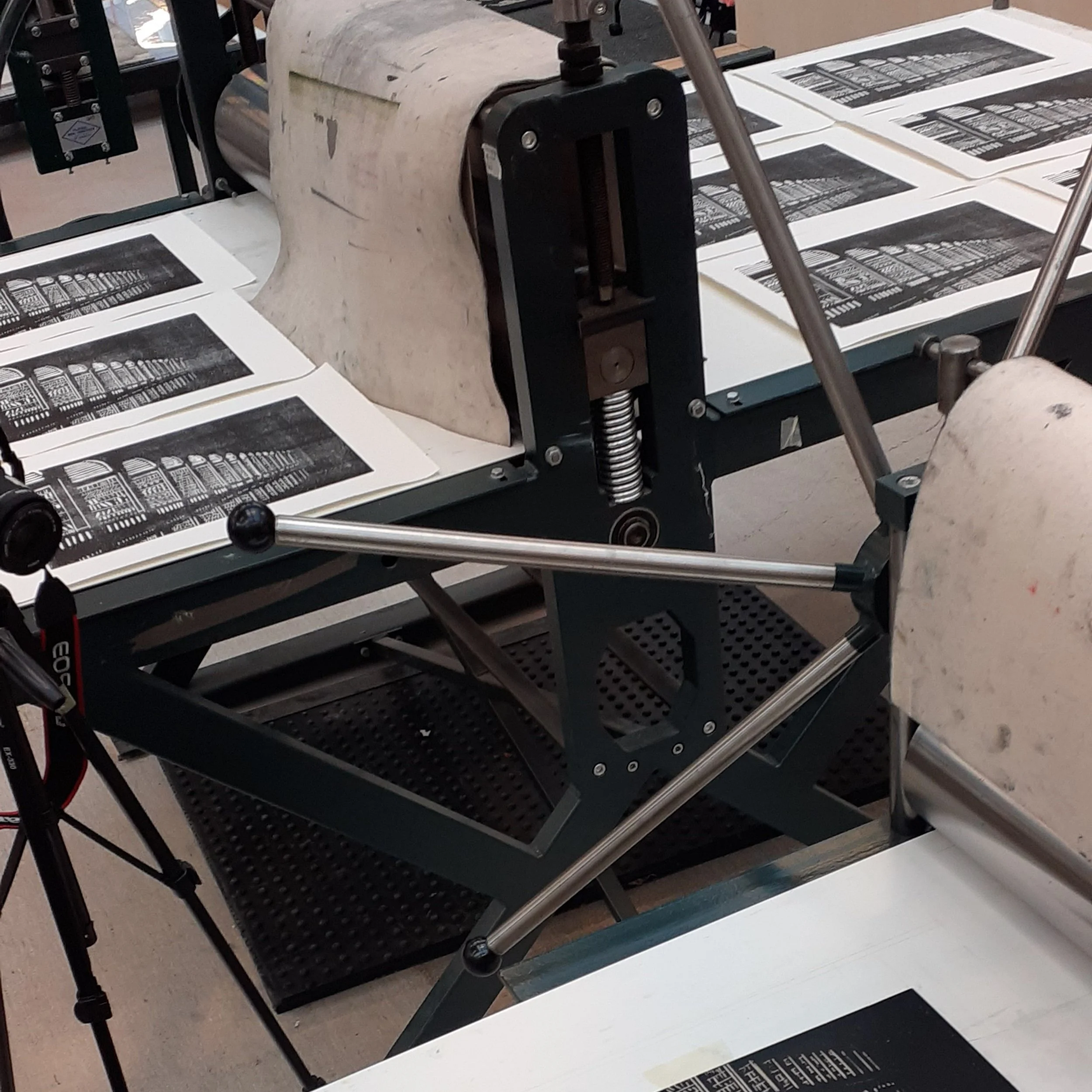 Printmaking press with sheets of black and white prints arranged on the press bed.