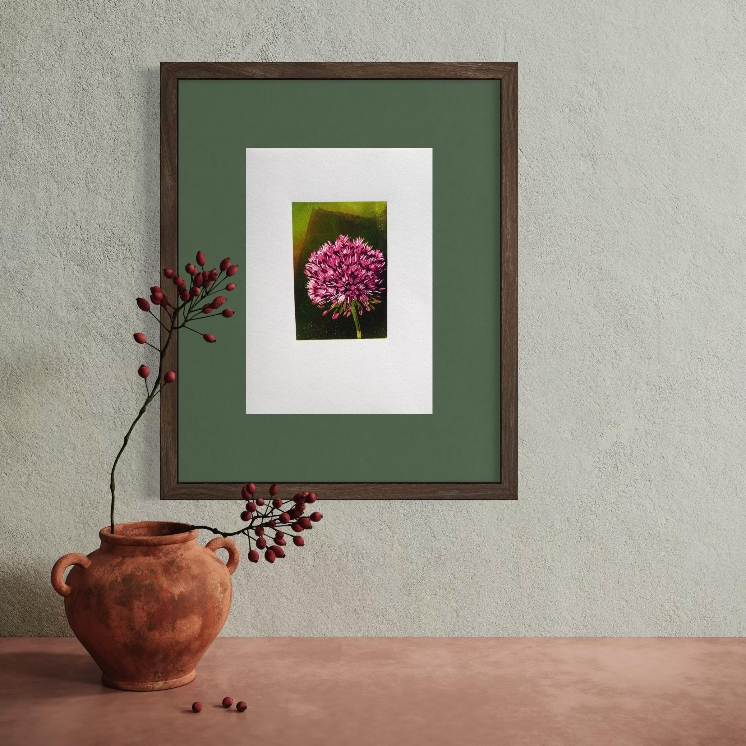 A ceramic vase with a round shape and two small handles on each side, filled with branches of red berries, is placed on a wooden surface. Behind it, a framed artwork highlights a pink flower against a green background, hanging on a beige textured wall.