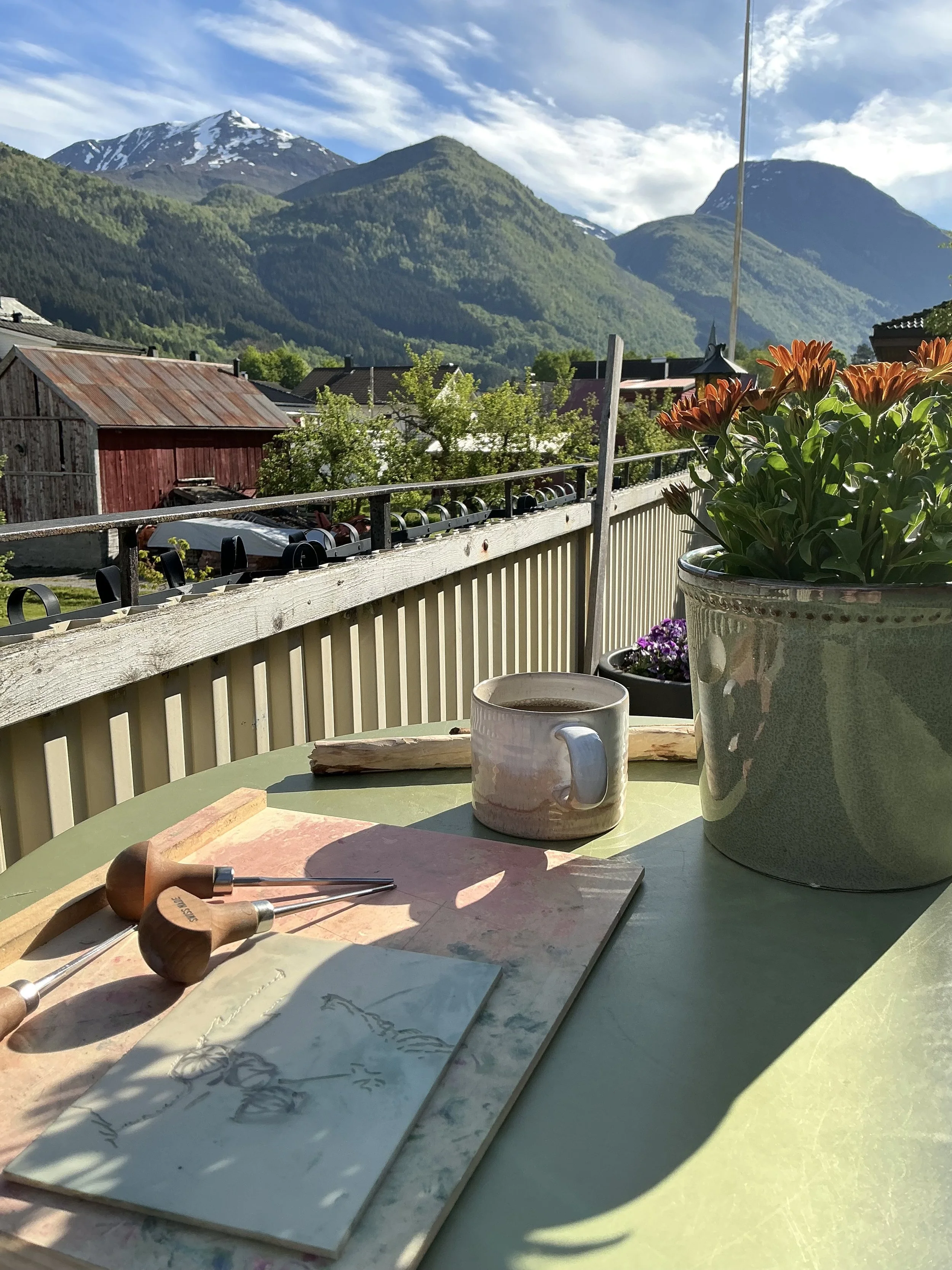 A table with woodworking tools, a mug, and sketches of trees. A large flowerpot with orange flowers and purple foliage is on a balcony, overlooking houses, green trees, and mountains in the background under a partly cloudy sky.