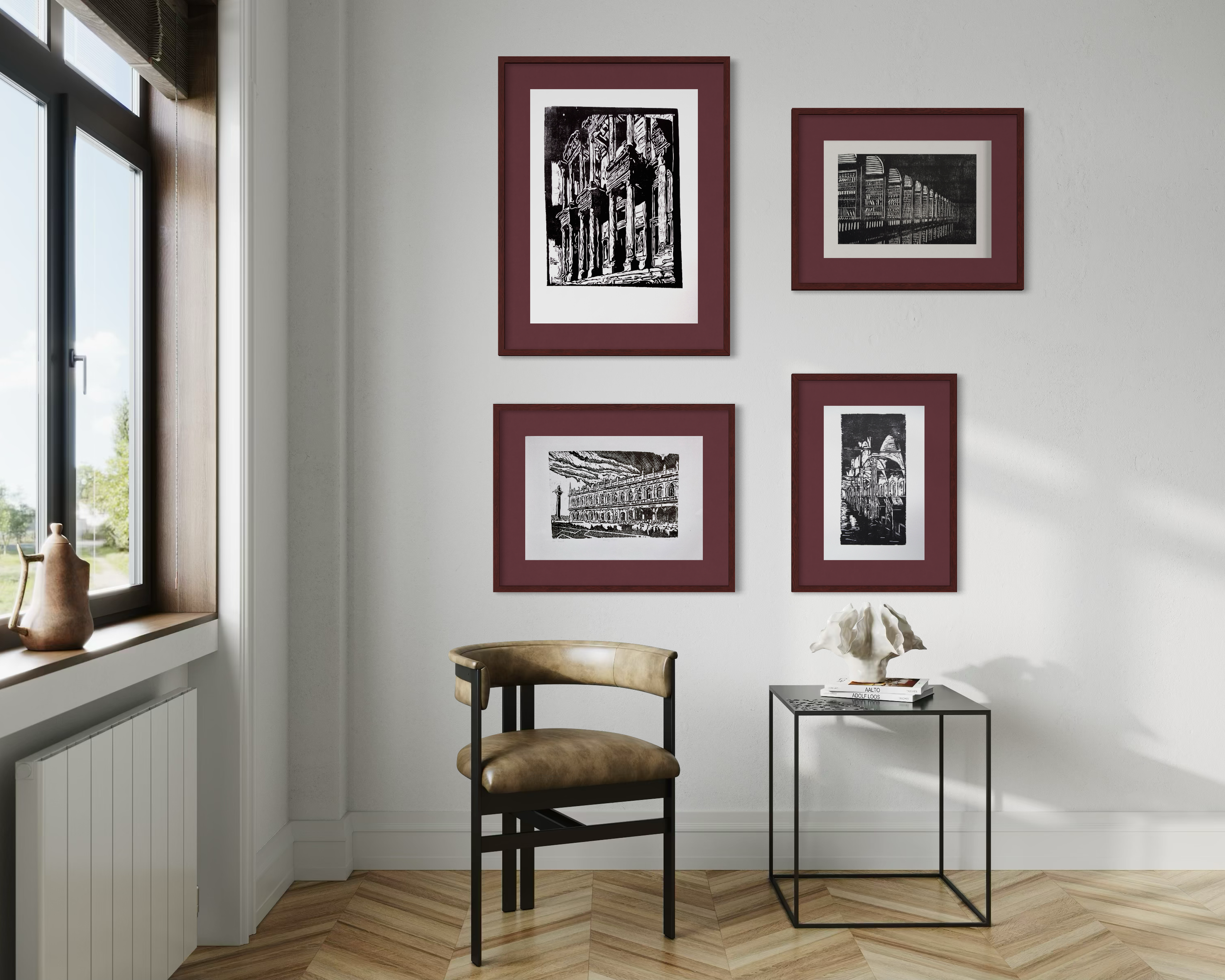 A corner of a room with a white wall featuring four framed black and white architectural prints. A window with wooden trim is on the left, allowing natural light. There is a small table with books and a decorative white sculpture, a beige chair, and a black metal side table.