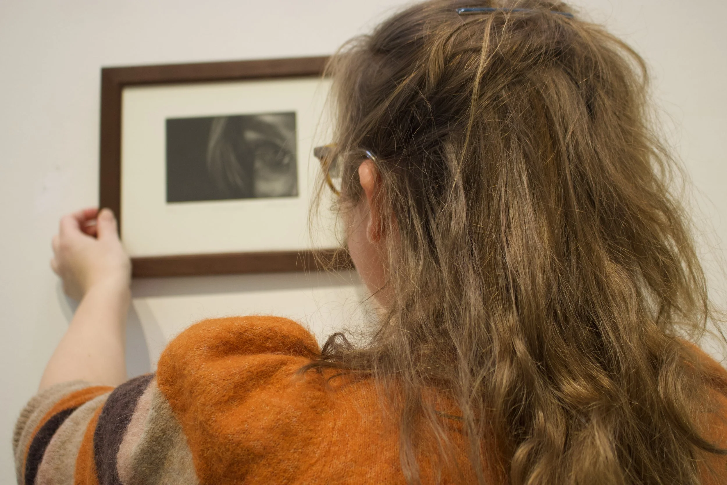 woman with brown hair, seen from the back, who hangs a framed mezzotint print on a white wall