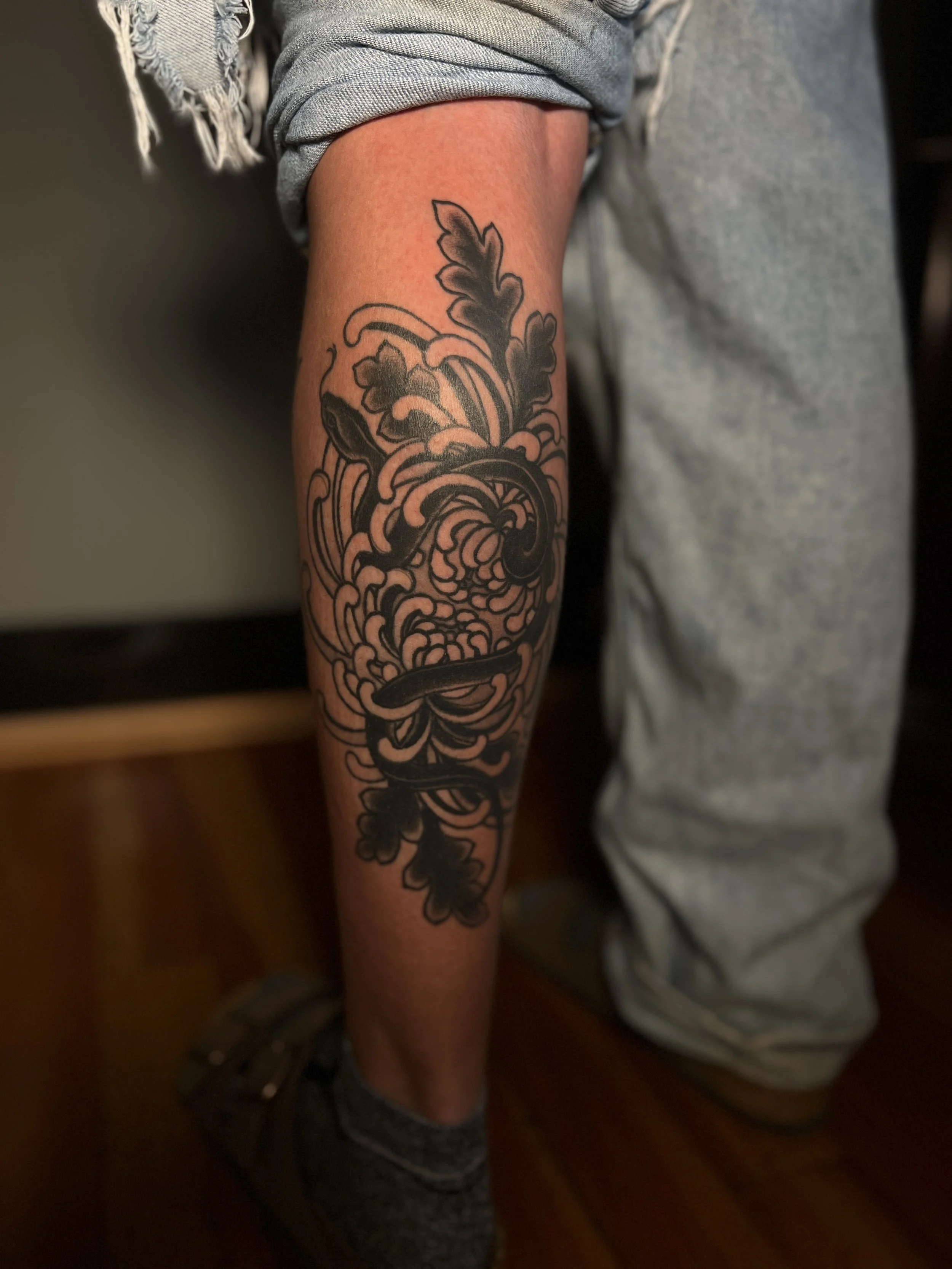 Trippy chrysanthemum snake leg tattoo with dark floral elements, designed and tattooed by Maya Wild at Sub Rosa Tattoo Studio in Grants Pass, Oregon. This unique piece highlights bold contrast and thick lines. Ideal for clients looking for dark flora