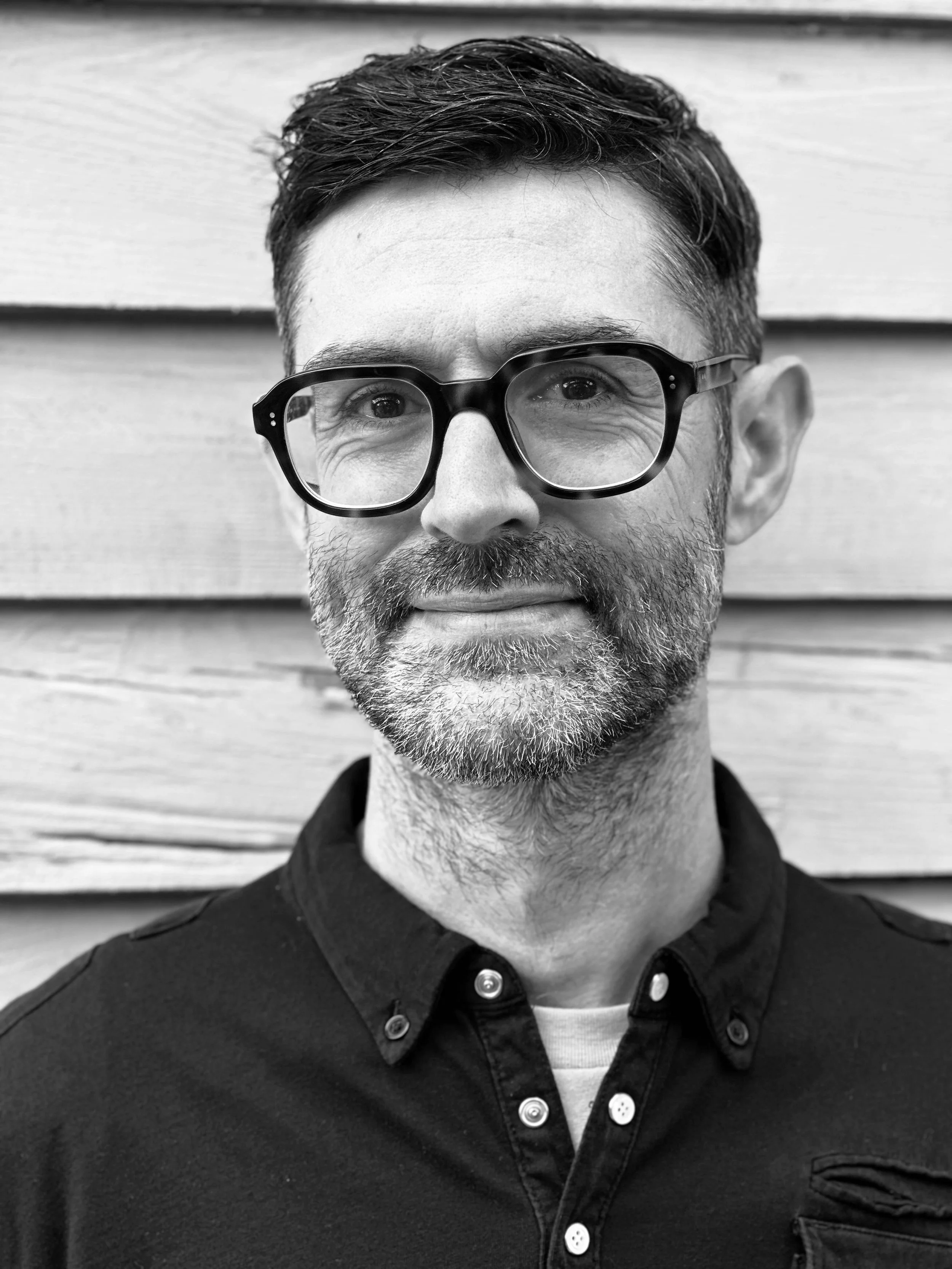 Black and white photo of a man with glasses, short dark hair, and a beard, standing in front of a wooden wall.