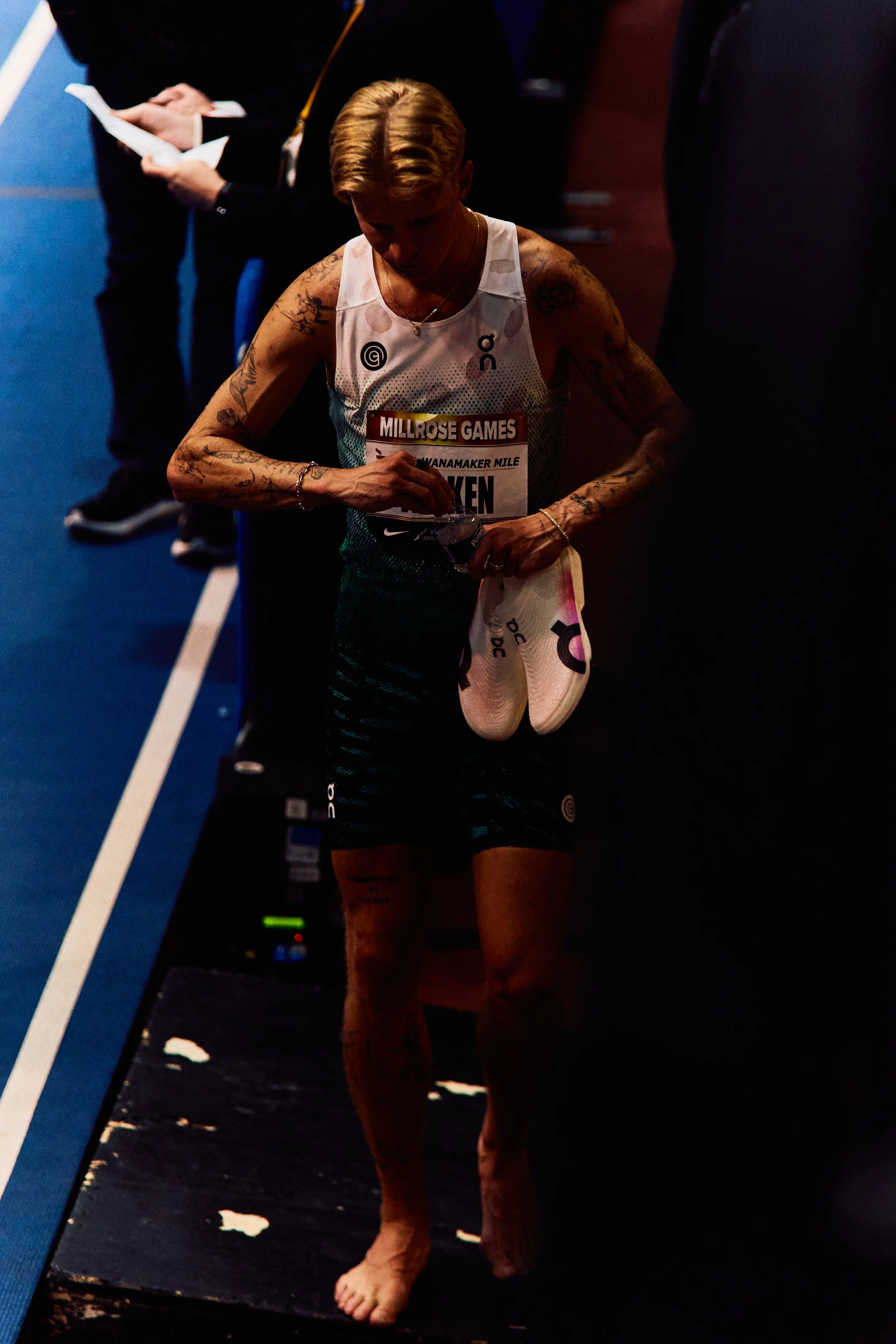 Female marathon runner with tattoos, wearing a white tank top and black shorts, holding her shoes and water bottle, walking barefoot on a dark indoor track after a race.