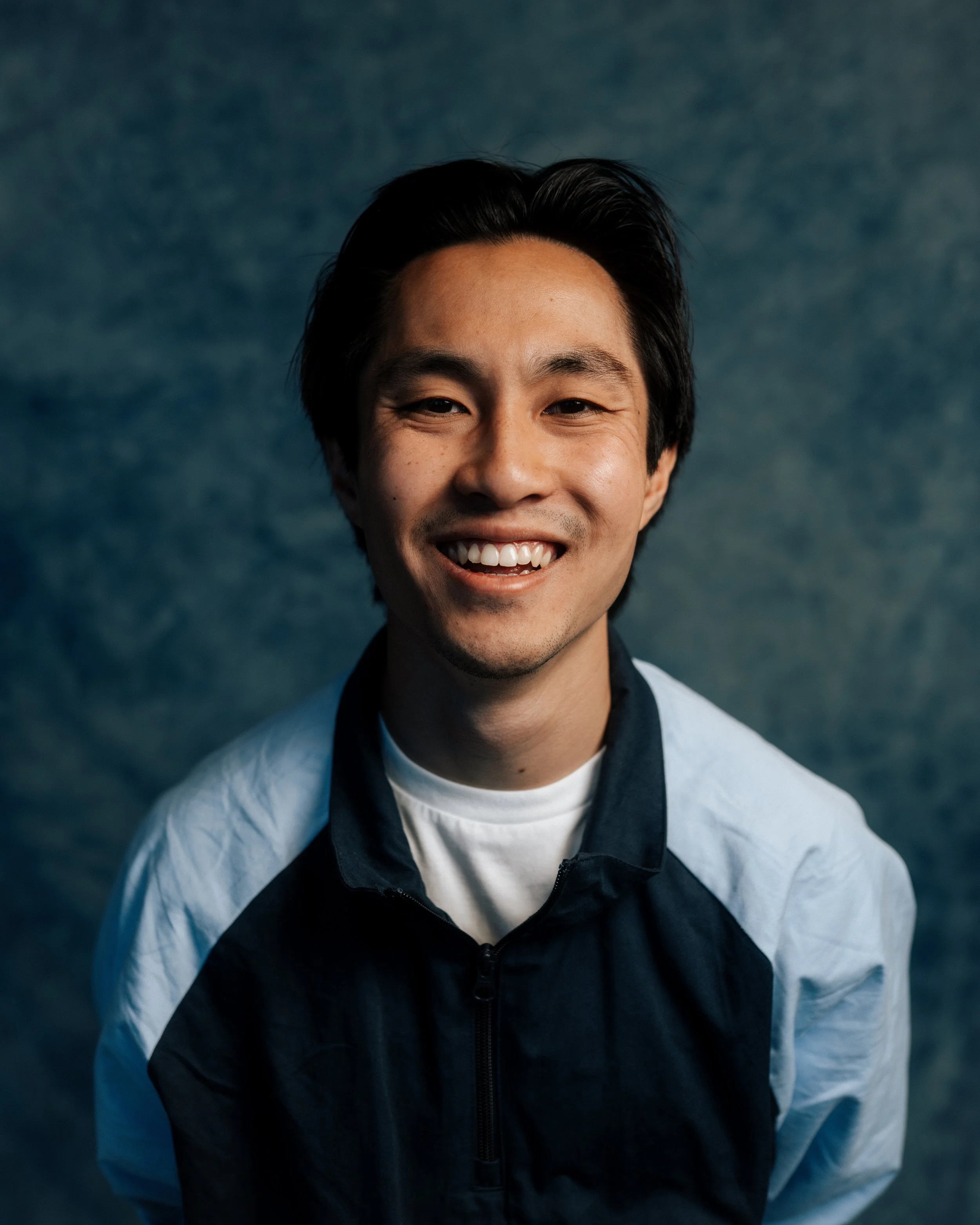 Close-up of a smiling young man with black hair, wearing a light blue jacket over a black shirt, against a dark, textured background.