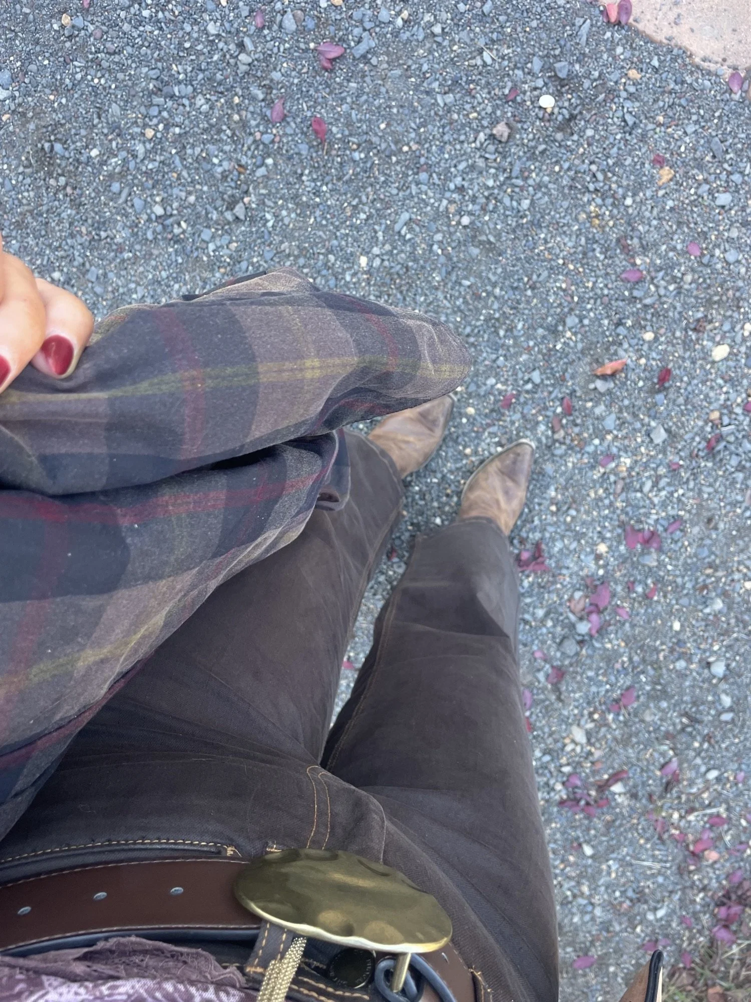 View from above of a person standing on a gravel ground with scattered pink flower petals. The person is wearing dark pants, brown shoes, a plaid shirt, and has a brown belt with a large brass buckle.