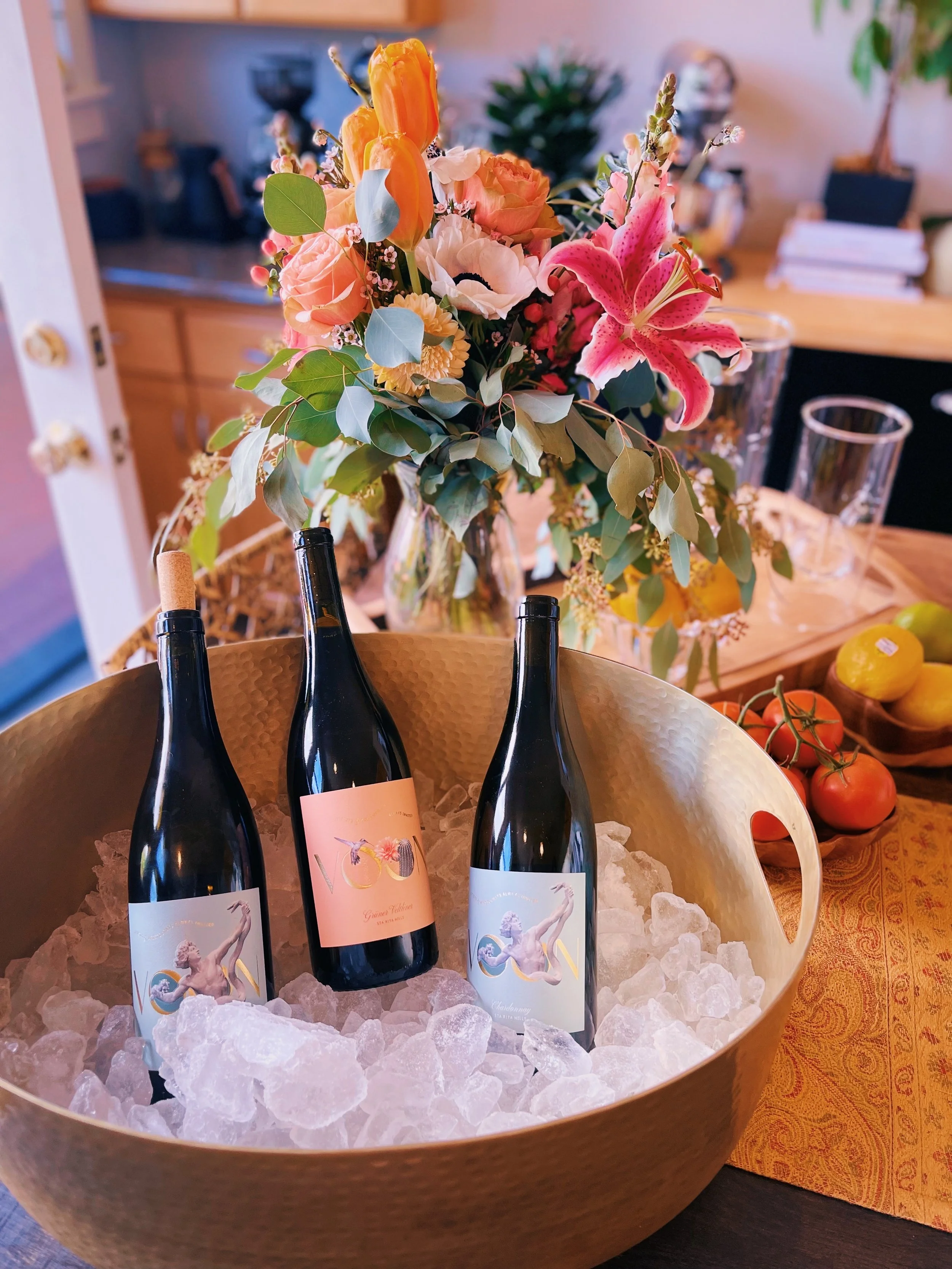 Three wine bottles in a gold container filled with ice, positioned on a wooden table with a yellow tablecloth. Behind them is a large bouquet of pink lilies, peach roses, and eucalyptus leaves on a wooden surface in a residential setting.
