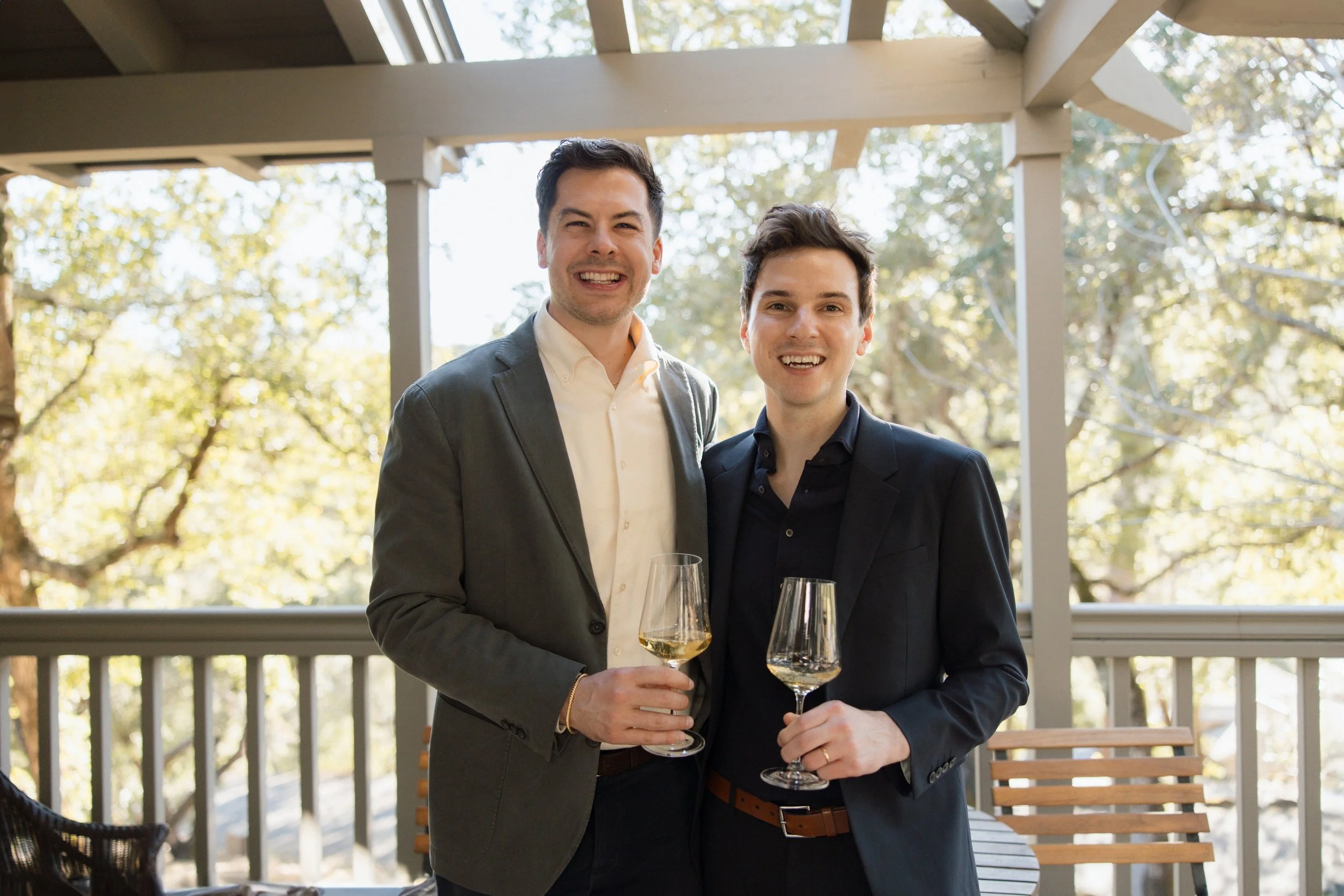 Evan and Cameron, owners of VOON Wines, dressed in suits holding wine glasses and smiling on a porch with trees in the background.
