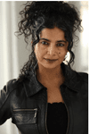 A woman wearing her long, dark, curly hair mostly up wears a black leather jacket and stands against a white background