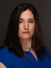 A woman with dark hair worn down and a serious face wears a blue top with beaded accents