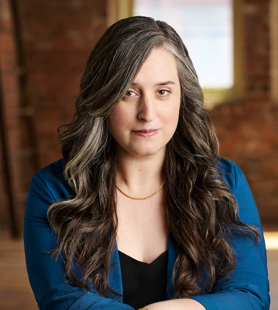 A woman with dark hair worn down and a serious face wears a blue blazer