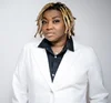A person with dark skin and gold-blonde braids wears a black and white suit agains a white background.