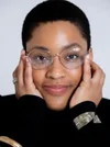 A Black woman with very short hair and wire-rimmed glasses with both hands on her face smiles