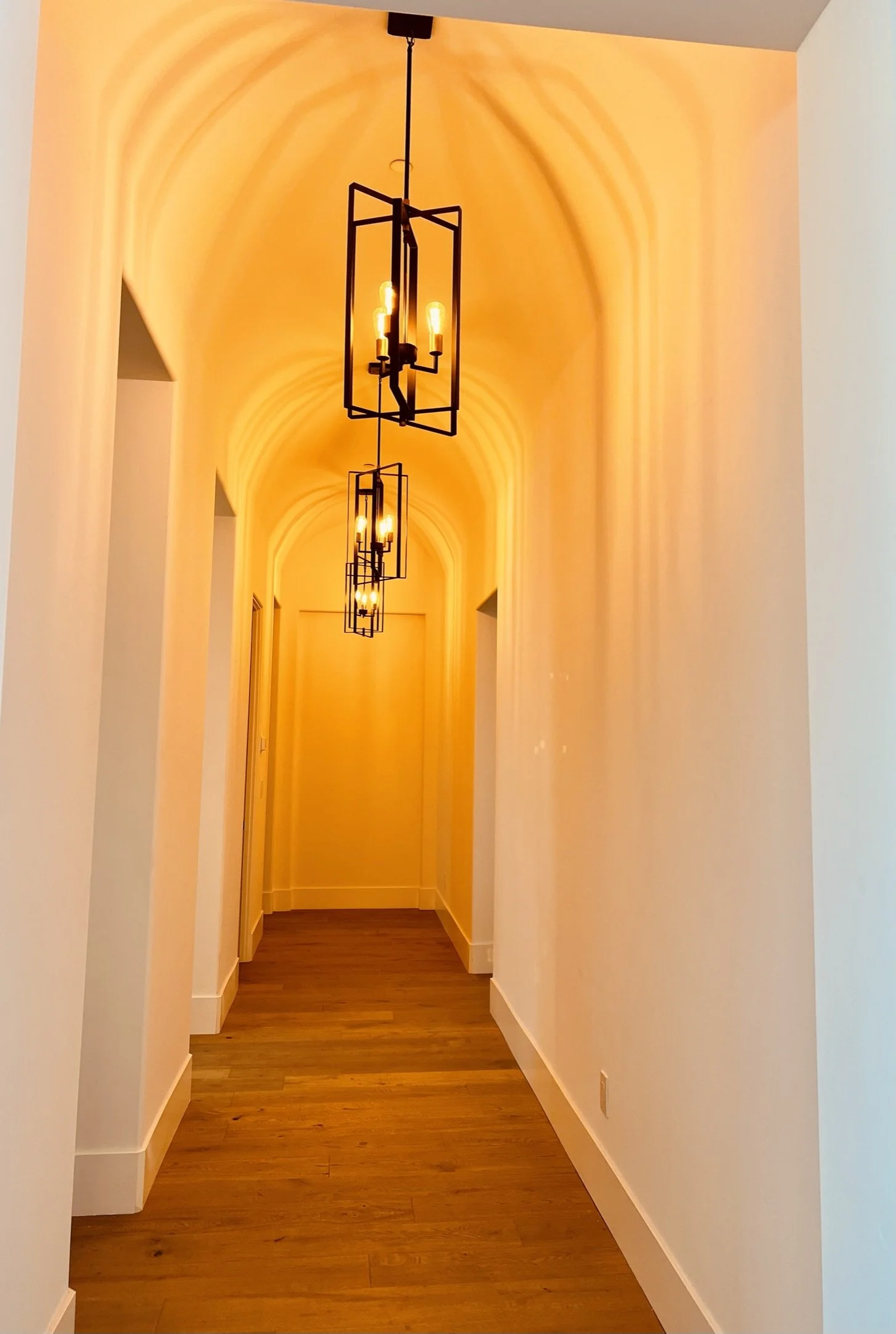 Long hallway with wooden floor and white walls, lit by modern black pendant chandeliers with exposed lightbulbs, and arched ceiling.