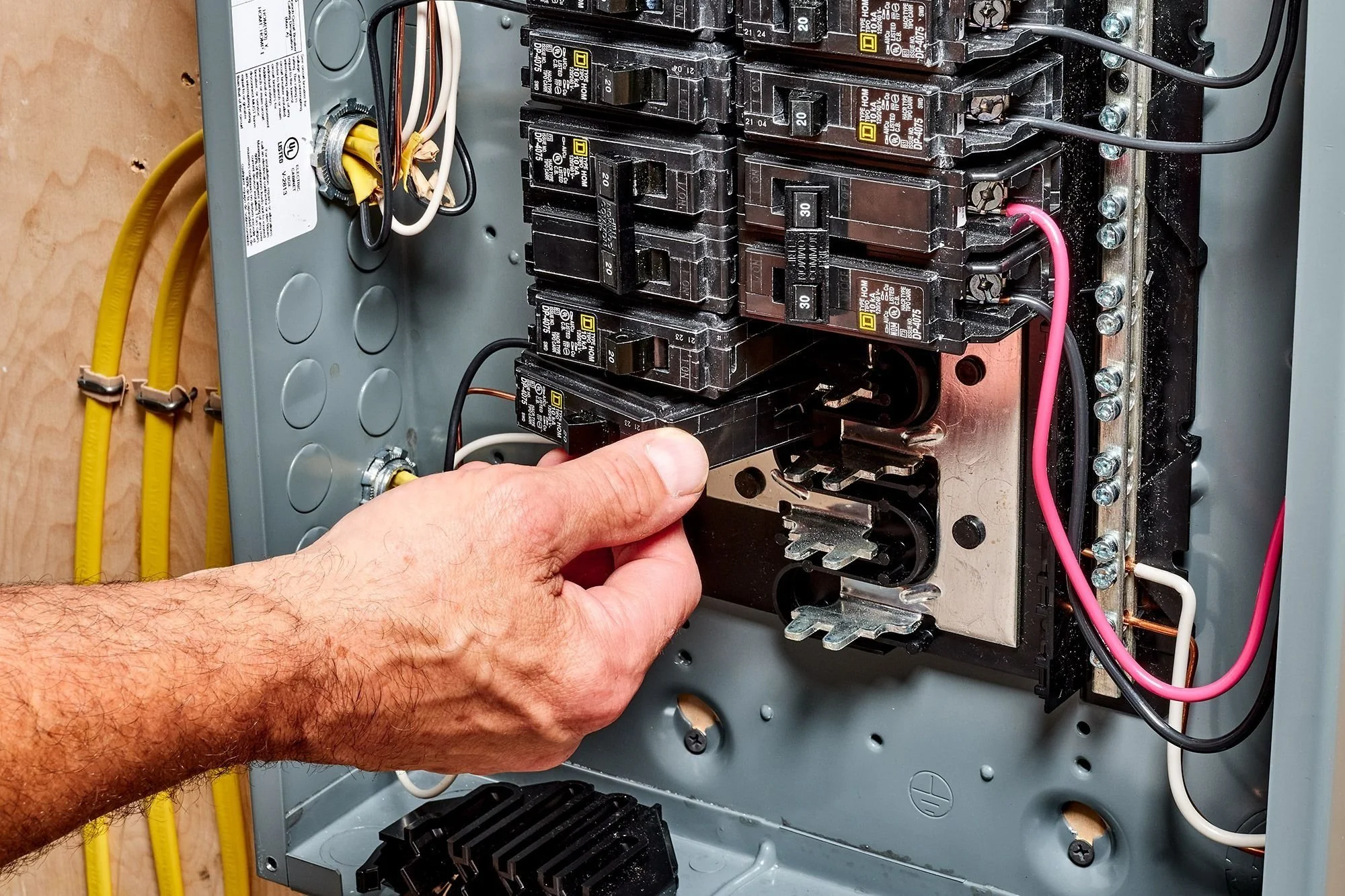 Close-up of a person's hand working on an open electrical circuit breaker panel, with a hand grasping a black circuit breaker.