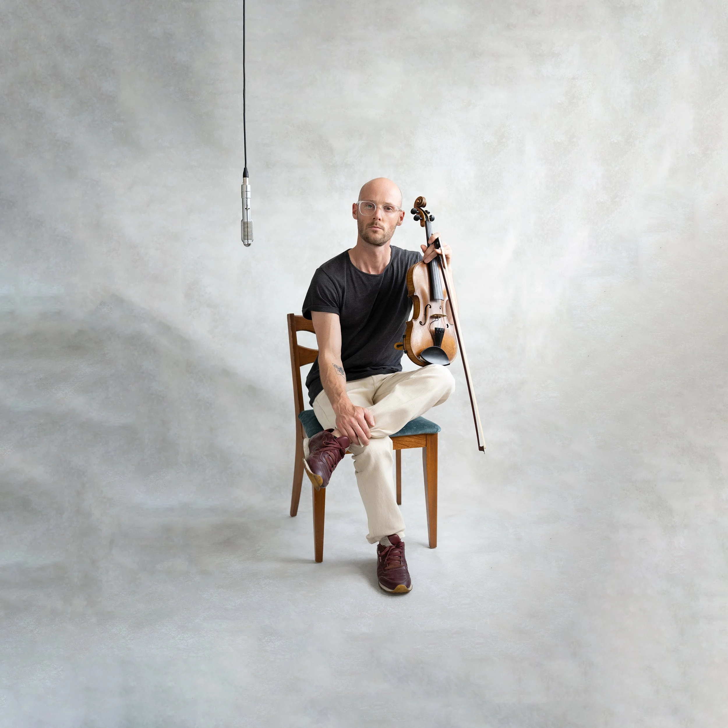 A man with glasses and a bald head holding a violin while sitting on a chair in front of a plain, light-colored wall with a microphone hanging from the ceiling.