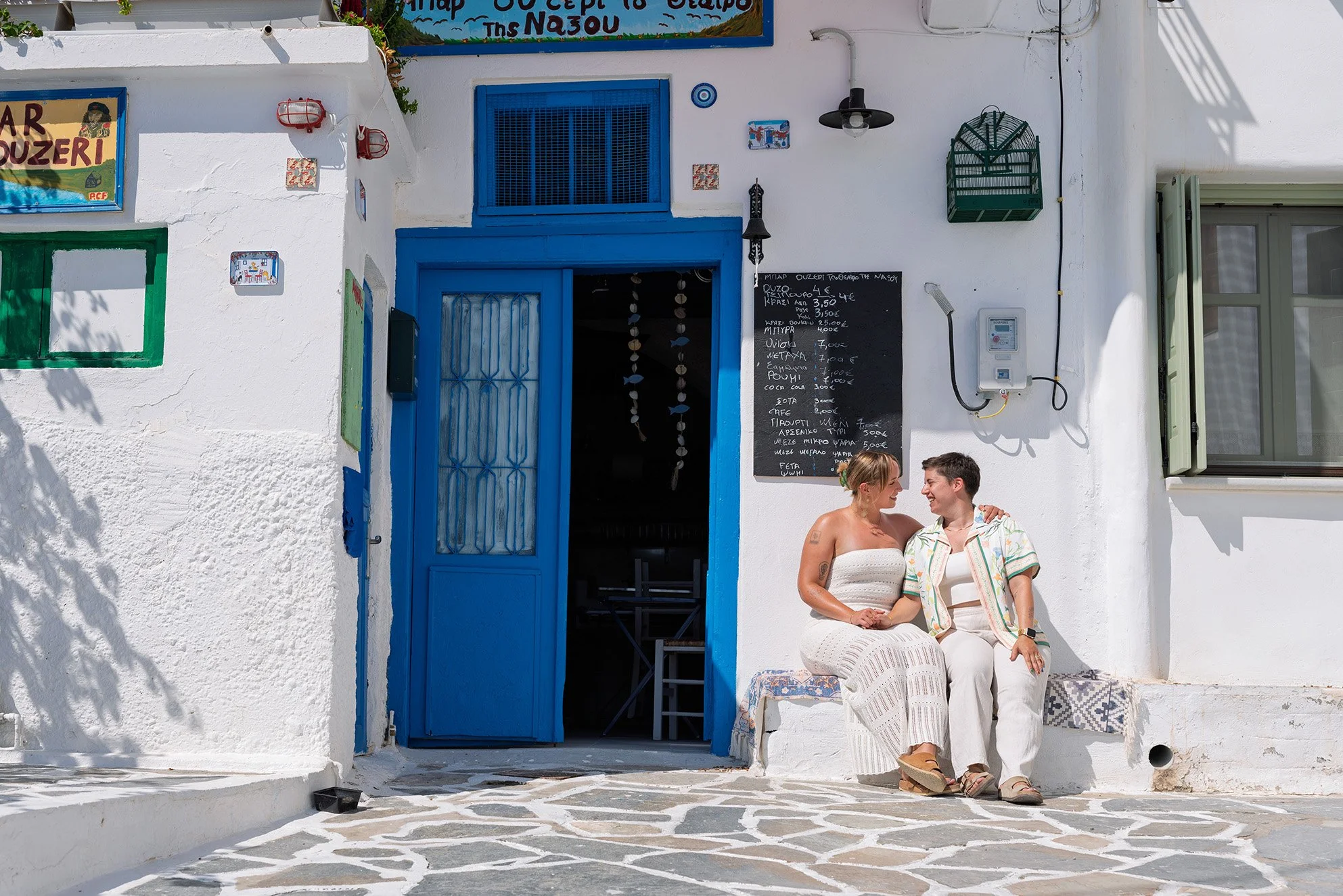 LGBTQ+ couple sharing a happy moment outside a traditional whitewashed taverna with blue doors in Naxos Old Town during a 2025 couple photoshoot by Naxos Photographer.