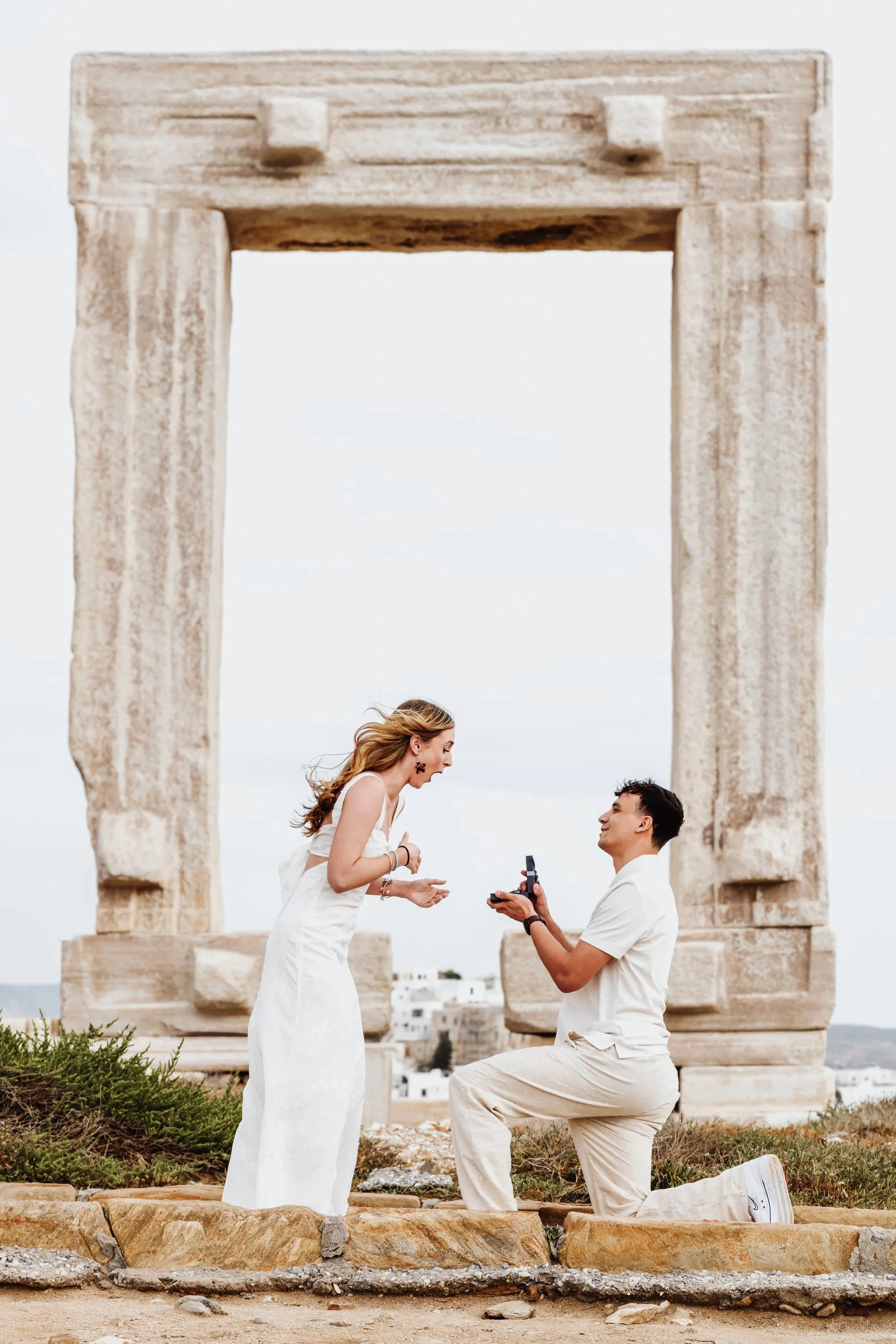 A man kneeling and proposing under the Portara in Naxos, with the woman reacting emotionally.