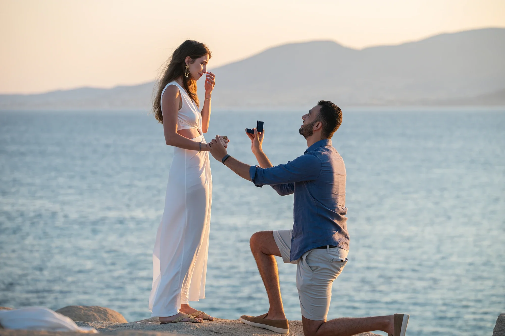 Surprise proposal photography in Naxos: a man kneeling with the ring by the sea at golden hour as his partner reacts with emotion.
